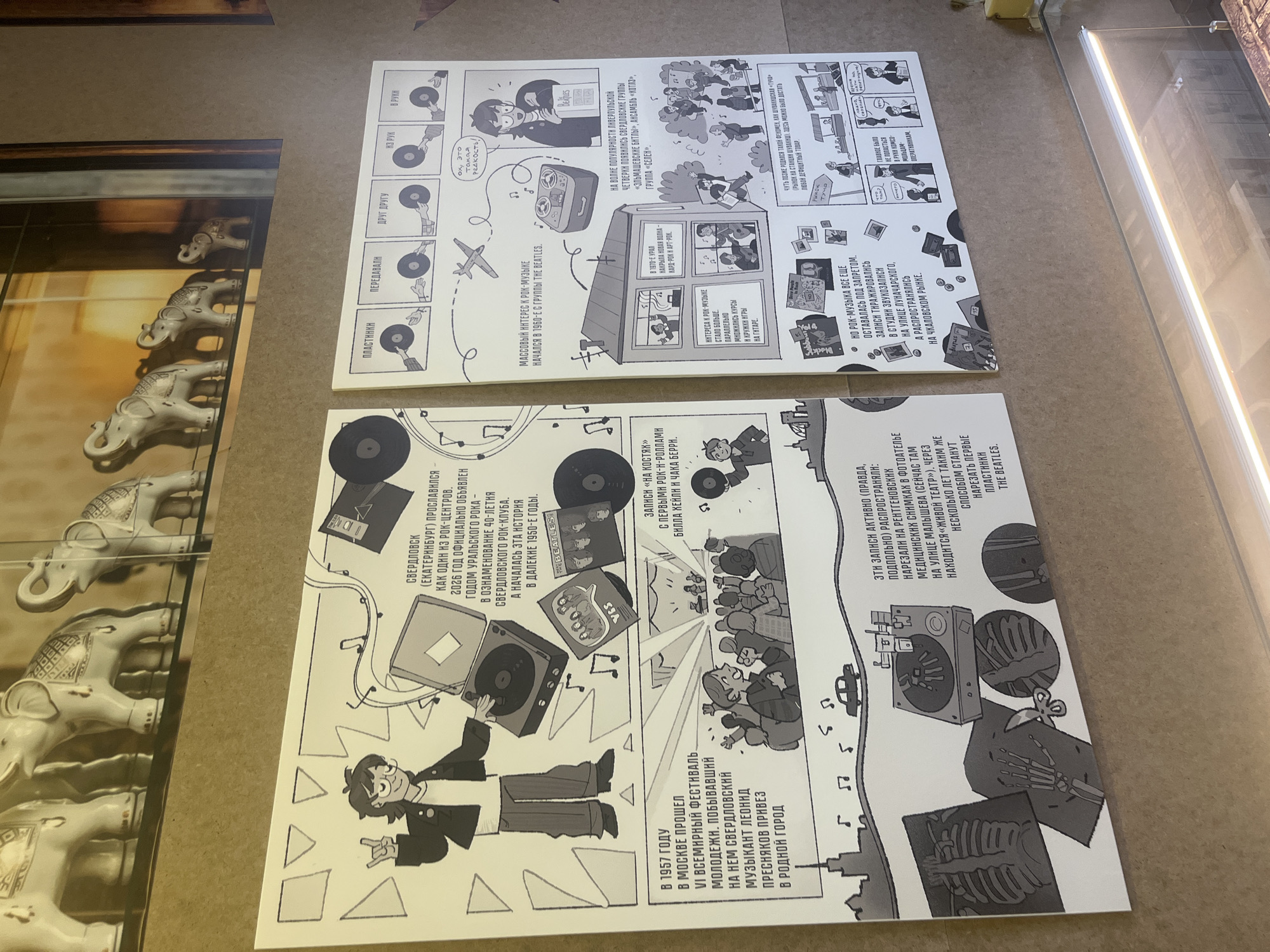The image shows two white posters displayed side by side. They appear to be informational panels with illustrations and text, likely about the history of rock music in Yekaterinburg, Russia.
The left poster features illustrations and text in Russian, discussing the history of rock music in Sverdlovsk (now Yekaterinburg). It mentions the 1957 World Festival of Youth in Moscow, which brought musical influence to the city. It also references the 40th anniversary of the Sverdlovsk Rock Club. The illustrations include a record player, vinyl records (one labeled "THE BEATLES"), and a crowd of people at a concert. At the bottom, there is a stylized cityscape of a town, with musical notes floating above the skyline. Towards the top right, the text mentions "records 'on bones'," alluding to illicit recordings made on X-ray films.
The right poster depicts different ways people interacted with vinyl records in Russia. It shows illustrations with captions describing different actions involving records, such as passing them to each other and playing them. It also discusses mass interest in rock music beginning in the 1960s with The Beatles. The poster mentions various Sverdlovsk bands and details about how rock music was still prohibited. The bottom section illustrates illicit recording and distribution methods, with "records on bones" made from x-ray films and being sold in a market.
In the background, there is a display case containing a collection of ceramic elephants. The posters are on a neutral-toned surface. The lighting appears to be artificial and somewhat bright.