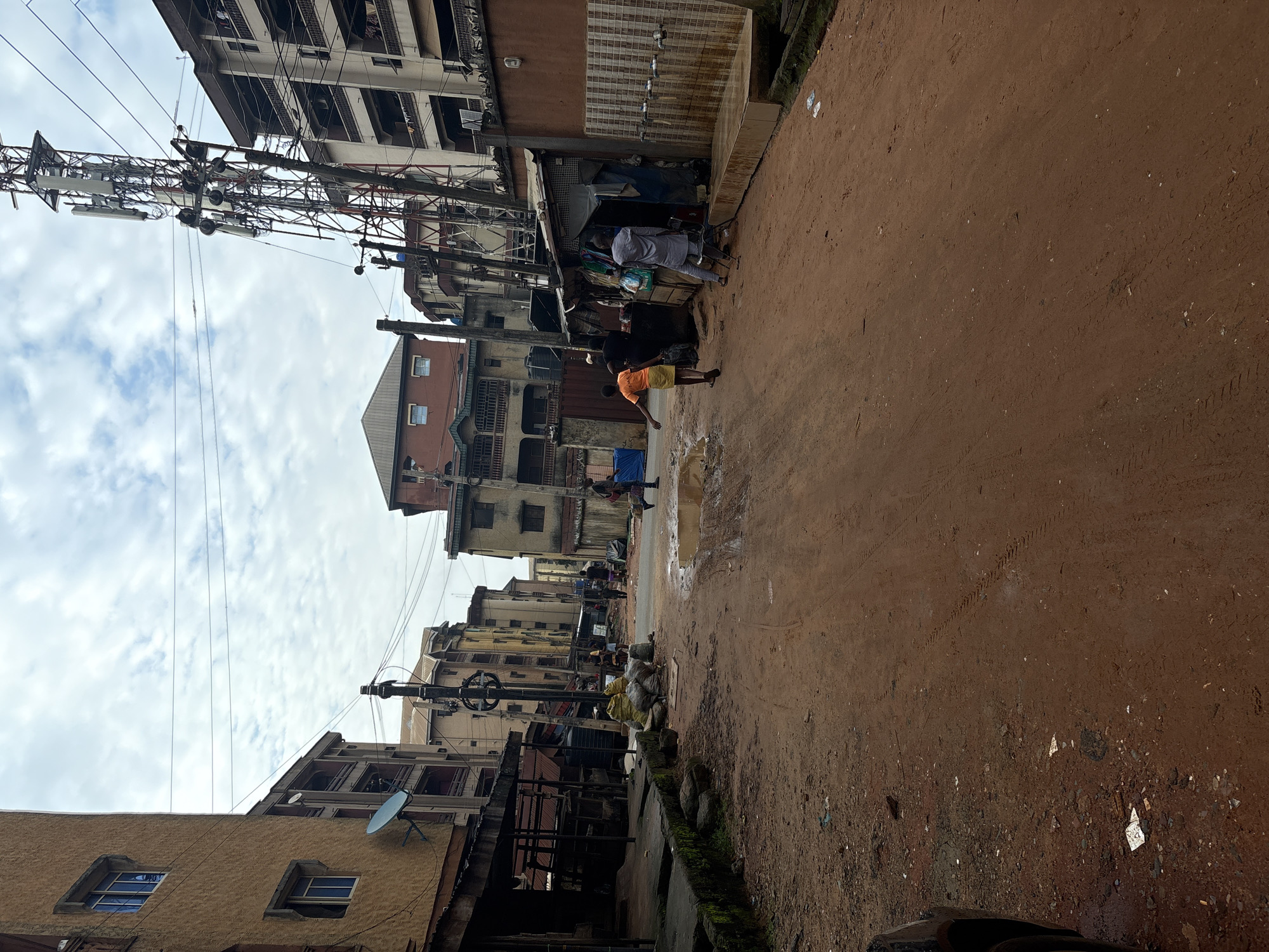 The image depicts a street scene in Onitsha, Nigeria, characterized by red dirt roads and multi-story buildings. In the foreground, the road is uneven with tire tracks and puddles of water, suggesting recent rainfall. To the left, a building with a textured yellow facade features several windows and a satellite dish. Adjacent to it is a covered market or workshop area with wooden supports and a corrugated metal roof.
In the middle ground, several people are visible. A young person in an orange shirt and yellow shorts walks away from the camera. Another individual, wearing a light-colored tunic and trousers, stands near a stall. Further down the street, other figures are walking or standing, some near a large communication tower that dominates the right side of the frame. This tower is surrounded by wires and poles.
The background consists of a row of buildings, some appearing to be residential apartments with balconies and many windows. The sky is overcast with scattered clouds. The overall impression is of a busy urban environment with elements of informal commerce and infrastructure.