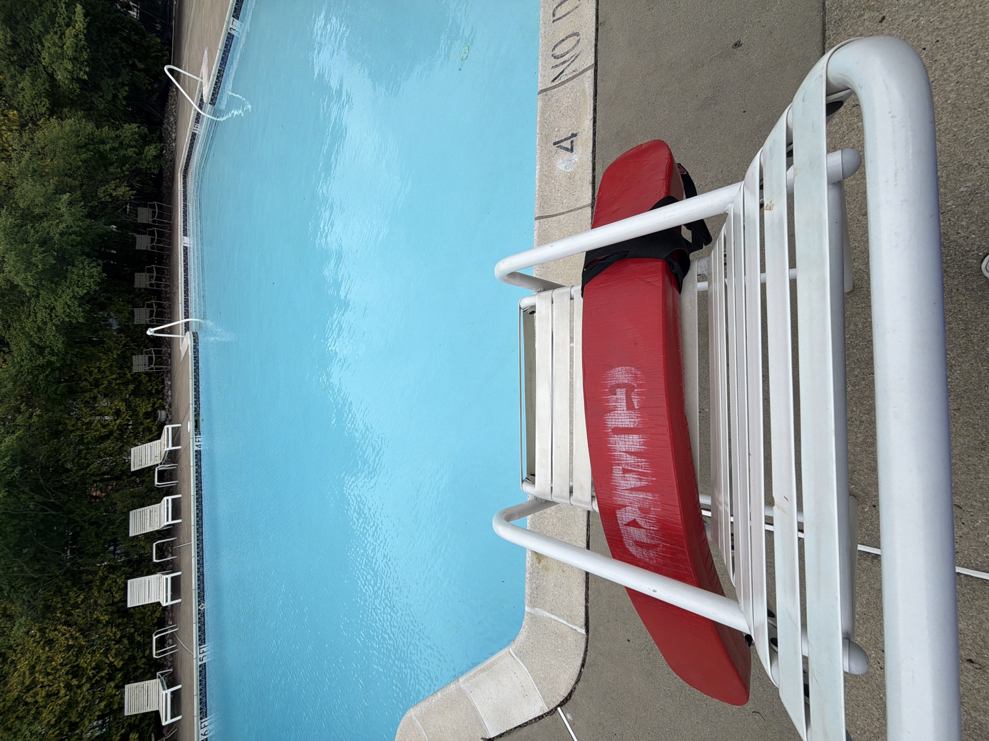 This is an outdoor scene at a swimming pool in Edison, New Jersey, USA. A red lifeguard rescue buoy is hanging on a white lifeguard chair at the edge of the pool. The pool water is clear and blue. Along one side of the pool are several white lounge chairs. Green trees and bushes line the area behind the lounge chairs. Pool depth markers are visible.
