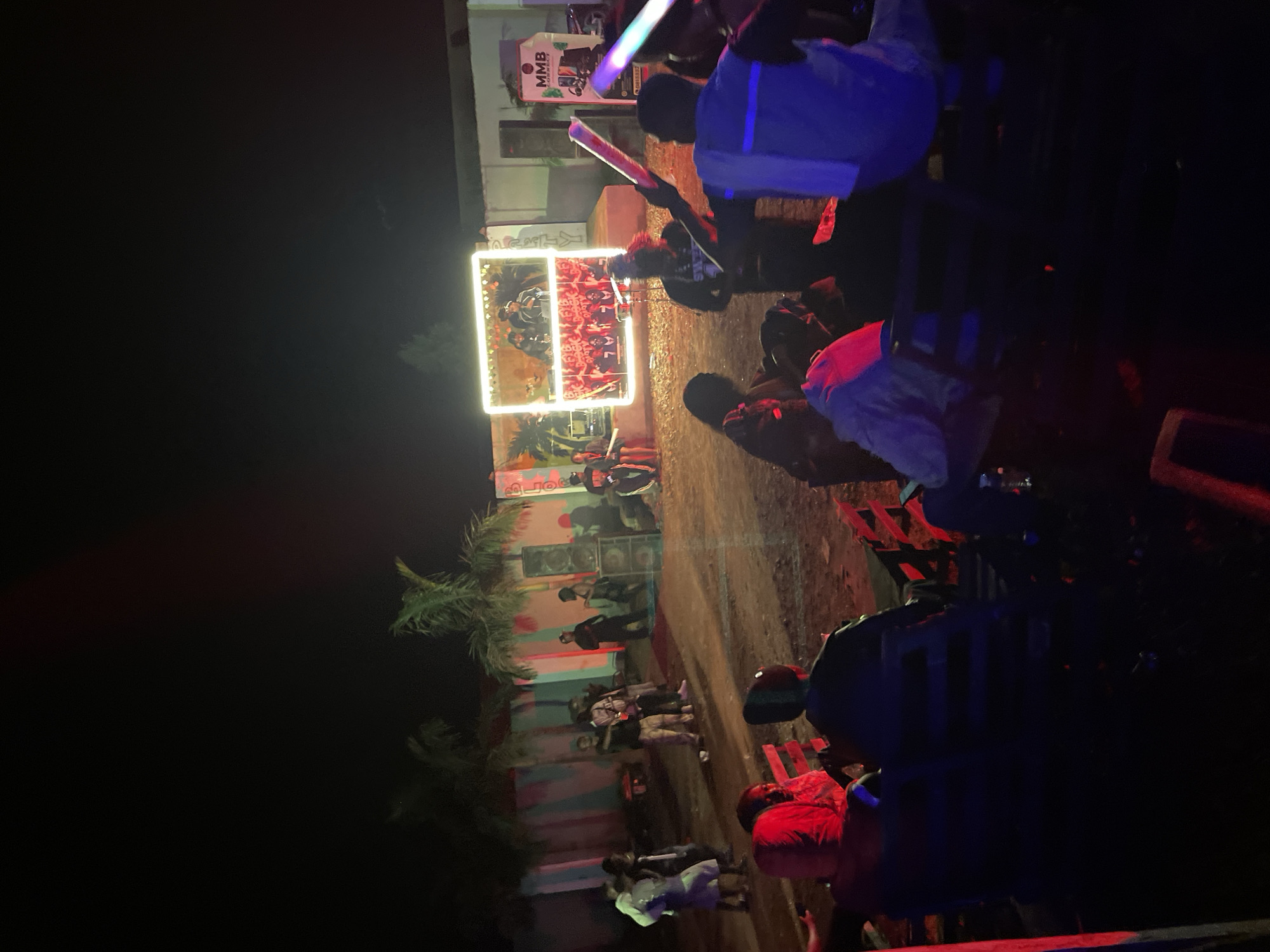 This image depicts an outdoor event at night in Durumi, Nigeria. A group of people are gathered, with some seated on chairs in the foreground and others standing and interacting closer to a stage area. The stage features a large illuminated screen displaying images of individuals, possibly performers or DJs. Several large speakers are visible, suggesting a musical performance or event. In the background, palm trees are present, and there's a building with colorful, possibly painted, walls. The overall atmosphere appears to be festive, with the use of colored lights adding to the ambiance. Some individuals are holding glowing sticks, indicating the presence of light entertainment or party favors.
