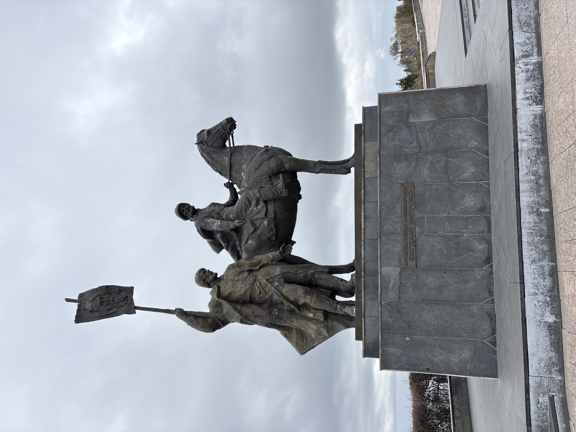 The image depicts a bronze monument in Ulyanovsk, Russia. A sculpture of a man on horseback is positioned atop a stone pedestal, with another man standing below holding a flag. The pedestal's surface contains inscription. The background reveals an overcast sky and hints of urban architecture.