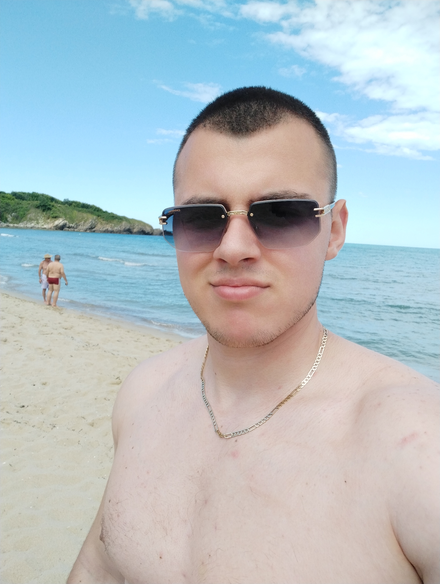 A close-up, selfie-style photograph shows a young man with short dark hair and a light beard, wearing dark-lensed sunglasses with gold frames and a gold chain around his neck. He is shirtless, exposing his chest and shoulder. The background consists of a sandy beach, calm blue ocean water, and a bright blue sky with scattered white clouds. In the distance, two men in bathing suits are walking along the shoreline. To the left, a lush green hillside with rocky outcrops rises from the water. The scene appears to be a daytime outdoor setting at a beach location.