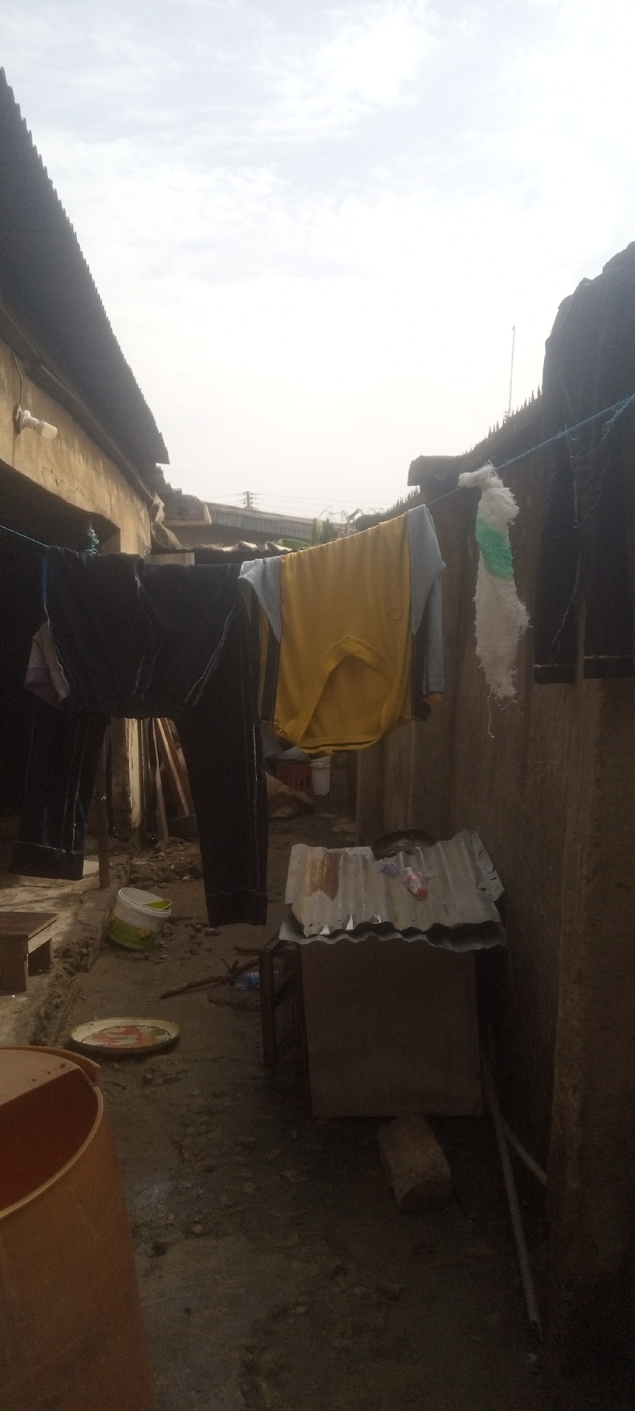 The image depicts a narrow alleyway in what is likely Sabon Tasha, Nigeria, given the user's prompt. A clothesline strung between two structures holds various articles of clothing, including dark pants, a yellow shirt, and other garments. The ground is uneven and appears to be dirt or packed earth. There are various objects scattered around, including a bucket, a metal plate, and a corrugated metal structure. The buildings on either side are constructed from what appears to be concrete and corrugated metal roofing. The sky is overcast. No people are directly visible in the frame. The general environment suggests a low-income, densely populated urban area.