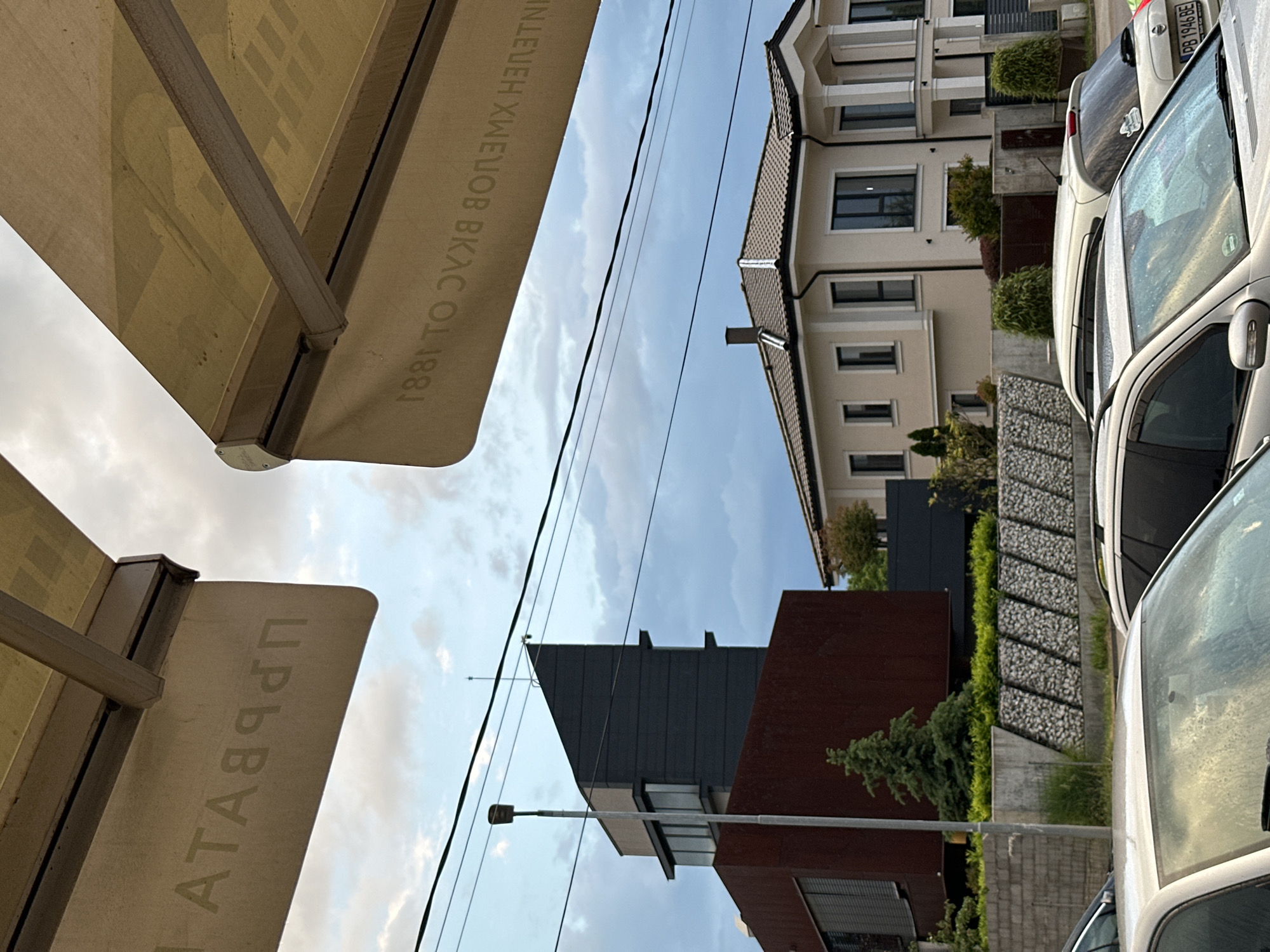 A low-angle view captures an urban scene featuring buildings, vehicles, and street-level elements. In the foreground, an awning with Cyrillic text is prominently displayed. A neighboring building with a modern architectural style features a mix of gray and brown materials. Further away, a traditional-style building with beige walls and dark-framed windows stands next to parked cars. The car closest to the viewer has a blurred windshield and a visible license plate. Power lines traverse the sky, which is partly cloudy. The scene is set in Plovdiv, Bulgaria.