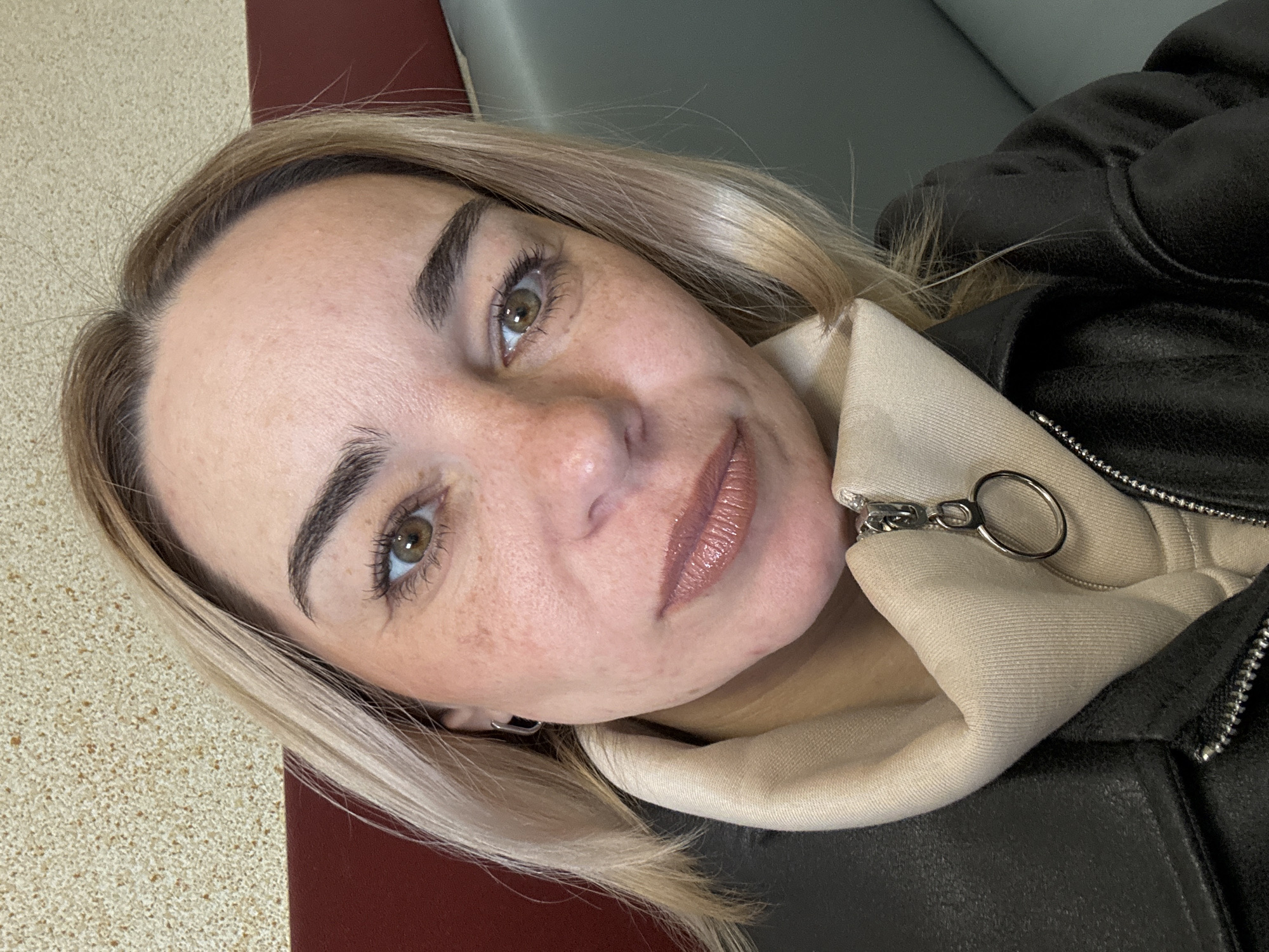 A close-up portrait depicts a woman with fair skin, light blonde hair, and hazel eyes. She is facing the camera at a slightly angled perspective, giving a soft smile. Freckles are visible on her face. She appears to be wearing a tan-colored hooded sweatshirt, partially obscured by a black leather jacket that is zipped up. The background is blurred, revealing a red surface on the left and a grayish-green area on the right, suggesting indoor seating or furnishings. There is no visible evidence to connect the scene with the city of Saint Petersburg, Russia. The location is otherwise indeterminate.