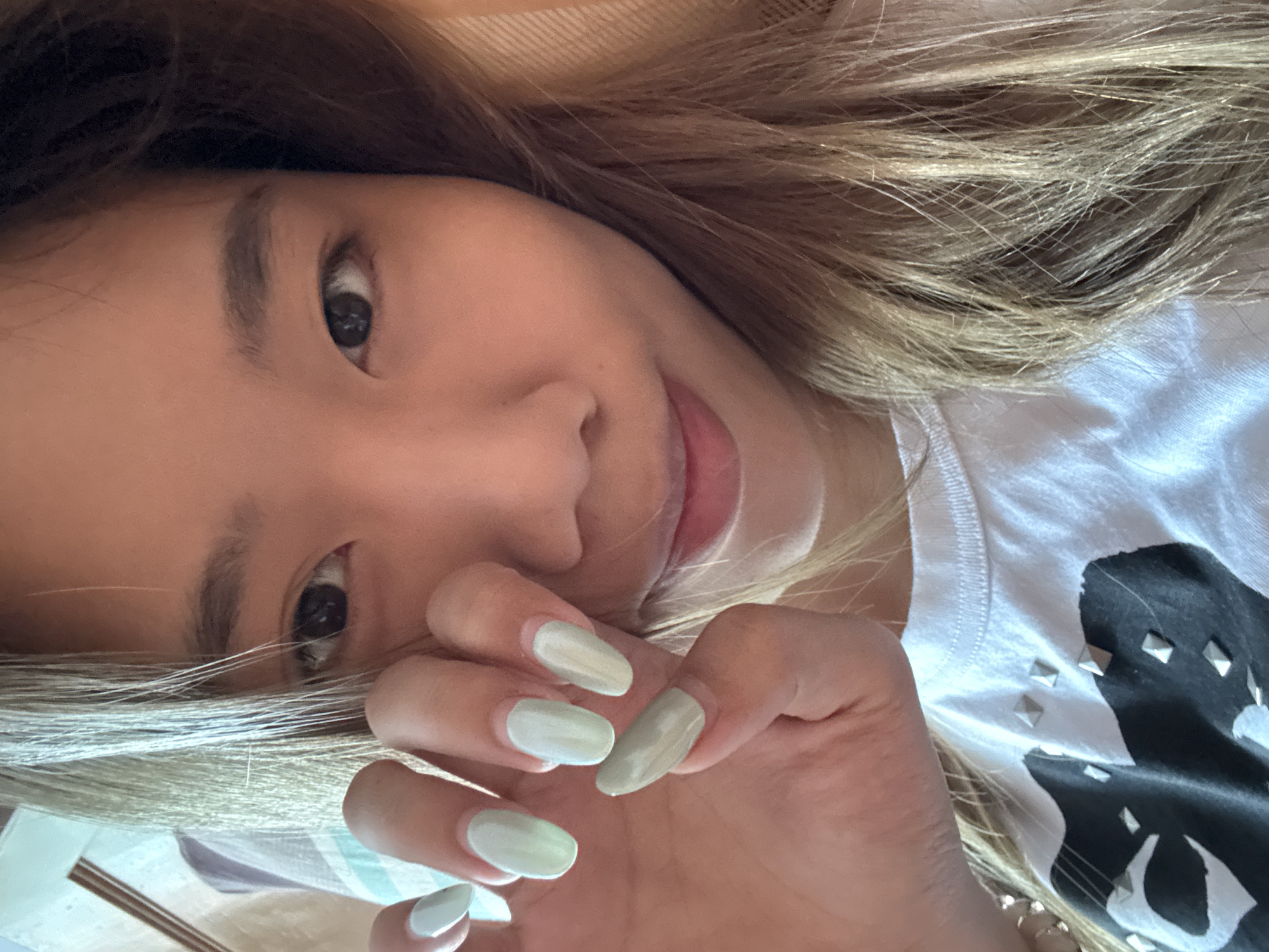 A close-up selfie shows a young East Asian woman from the chest up. She has light brown hair that frames her face and falls over her shoulders. Her right hand is raised to her face, with her fingers curved, revealing light green nail polish. She is wearing a white t-shirt with a black graphic design embellished with silver studs. The background is out of focus, but appears to be an indoor setting with a patterned fabric. No specific location in Guandu, China, can be identified.
