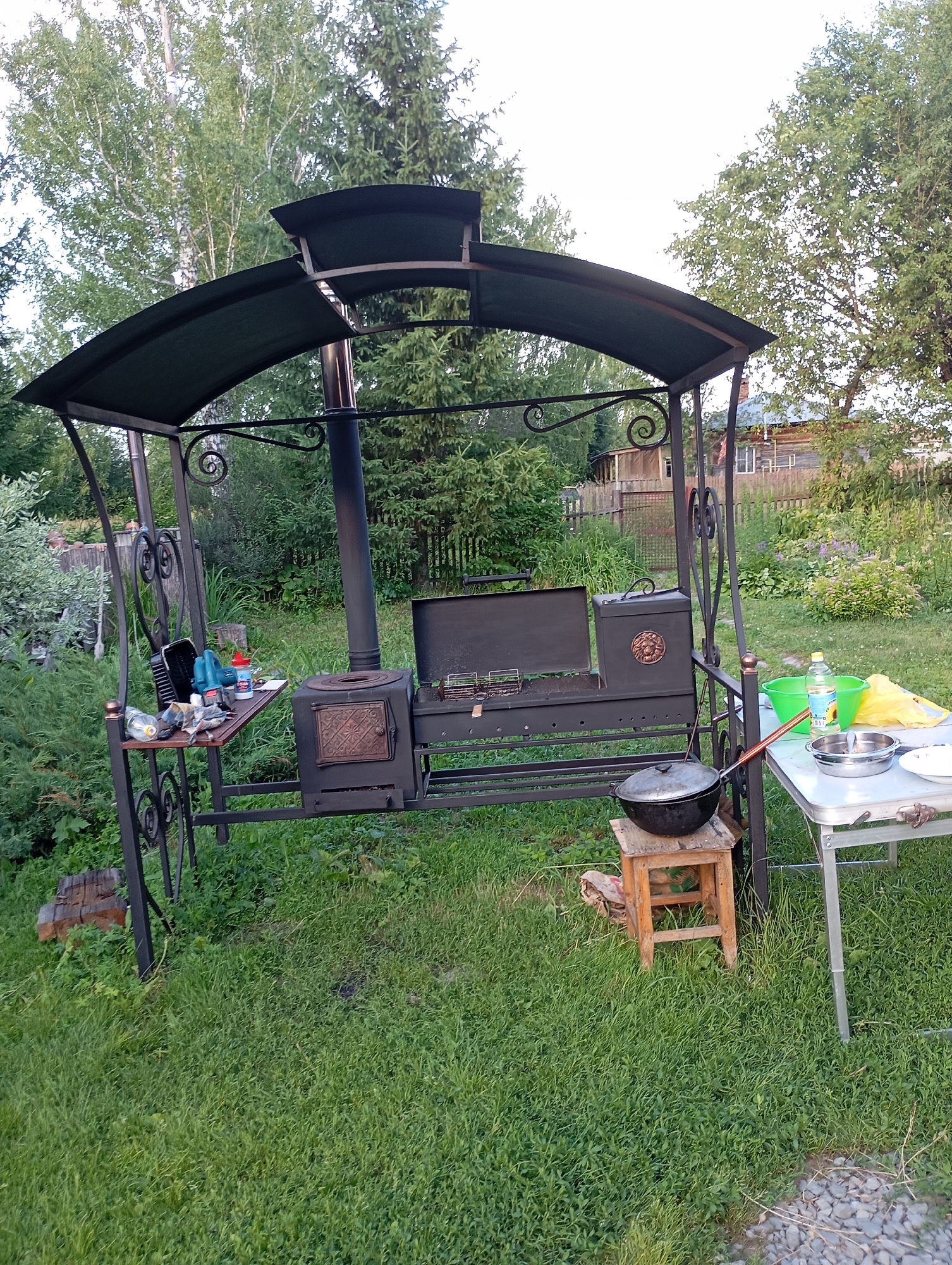 In Likhonikha, Russia, a backyard scene unfolds with a metal gazebo featuring a dark green canopy over a BBQ setup. The BBQ unit has a primary grill, a smaller oven, and a tall chimney. To the left, a small metal shelf attached to the gazebo holds various items, including bottles, a black container, and what appears to be a tool. A large black pot on a wooden stool sits in front of the BBQ. To the right, a folding table displays a green plastic basin, a bottle of oil, and two metal bowls. The gazebo is surrounded by lush green grass and vegetation, with trees and a wooden fence in the background. A two-story wooden house is visible behind the fence. The sky is overcast.