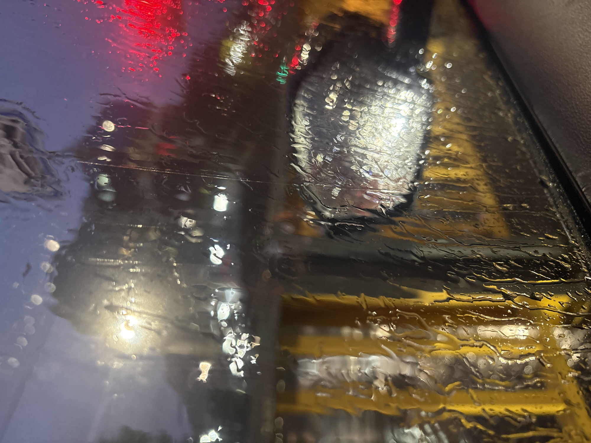 The image appears to be taken from inside a vehicle on a rainy night, possibly in Minsk, Belarus. Raindrops are heavily present on the windshield, distorting the view of the outside. Streetlights and traffic lights are visible, blurred and diffused by the rain. There is yellow infrastructure in the image as well. The general location is suggested by the urban environment and the language of the request.