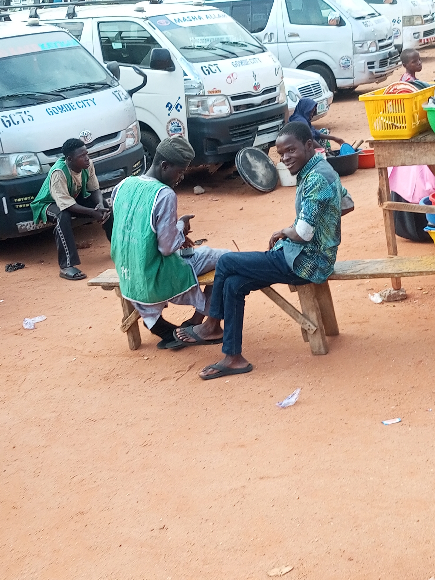The image shows a street scene in Gombe, Nigeria, with several vehicles and people. Two white vans, possibly minibusses, are parked side-by-side. One van has "GCT GOMBE CITY" and "173" visible on its side. The other van has "MASHA ALLAH" written on its windshield. A young boy in a pink shirt is visible near a yellow basket filled with dishes and a green container.
In the foreground, three men are present. One man, wearing a green vest over a light brown shirt and black pants, is sitting on the side of the silver van. Next to him, another man in a green vest over a grey garment and grey pants is seated on a wooden bench. He appears to be engaged in a conversation or activity with the third man, who is sitting on the same bench to his right. This third man is wearing a blue and green patterned shirt and dark blue jeans. His legs are crossed, and he is wearing dark sandals. The ground is covered in reddish-brown dirt.
A wooden bench with two people sitting on it is a prominent feature. A yellow plastic basket, likely containing kitchenware, is on the right side of the image, along with other containers. The scene appears to be outdoors, possibly a marketplace or a transportation hub, given the presence of the vans and the arrangement of goods. The lighting suggests it is daytime, likely mid-morning or afternoon. The overall atmosphere seems casual, with people gathered and interacting.