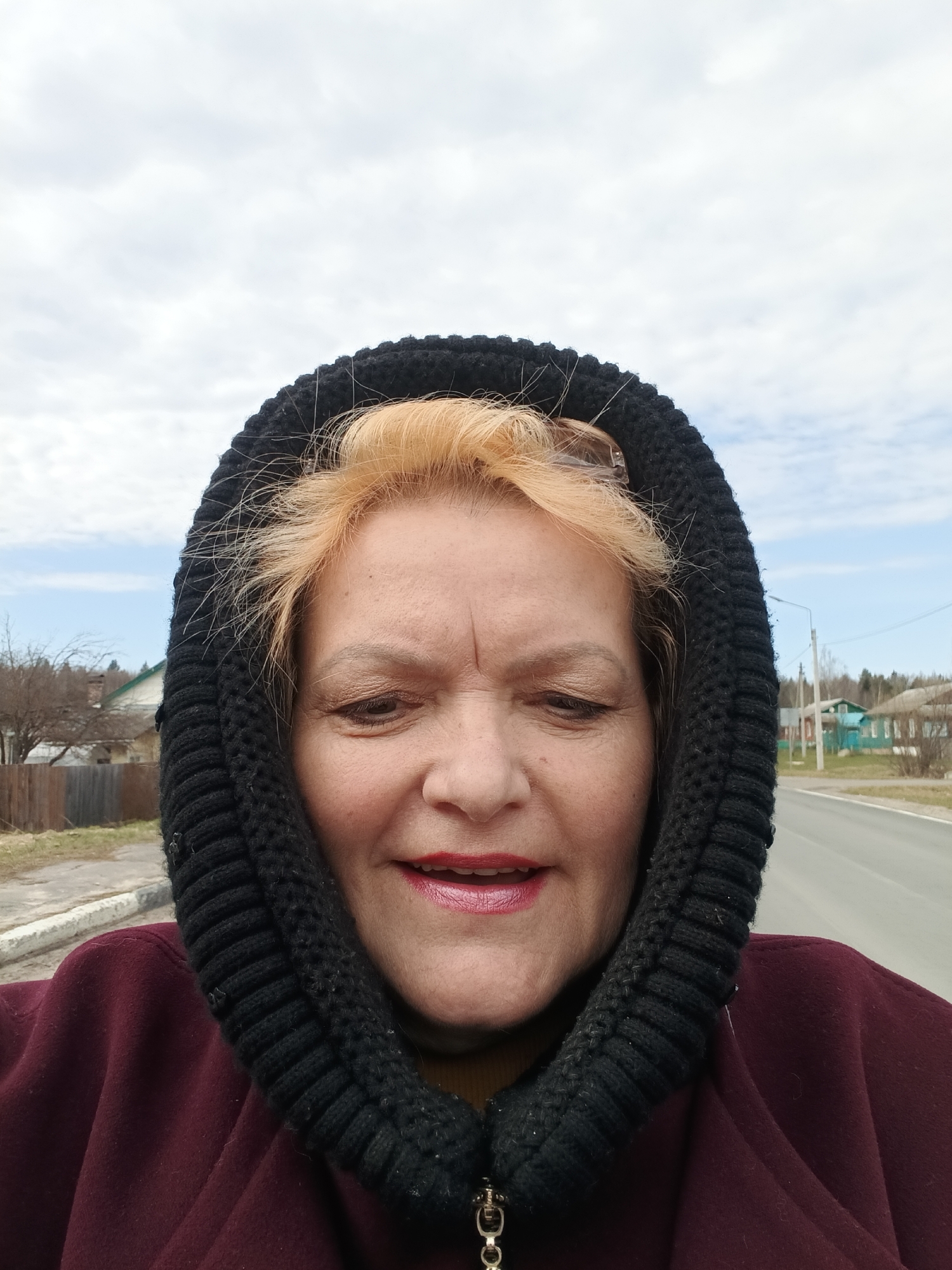 A medium shot captures a woman outdoors, with a black hooded garment framing her face. She has light orange hair and is wearing red lipstick. Her expression is one of slight amusement or happiness.
The background indicates an urban setting, possibly on the outskirts of a town. A paved road stretches into the distance, and on either side of the road are simple, likely residential buildings. The presence of power lines or poles is evident. The sky is overcast with patches of blue.
Based on the environment visible in the picture, it's possible this photograph was taken in Красный Маяк, Russia.