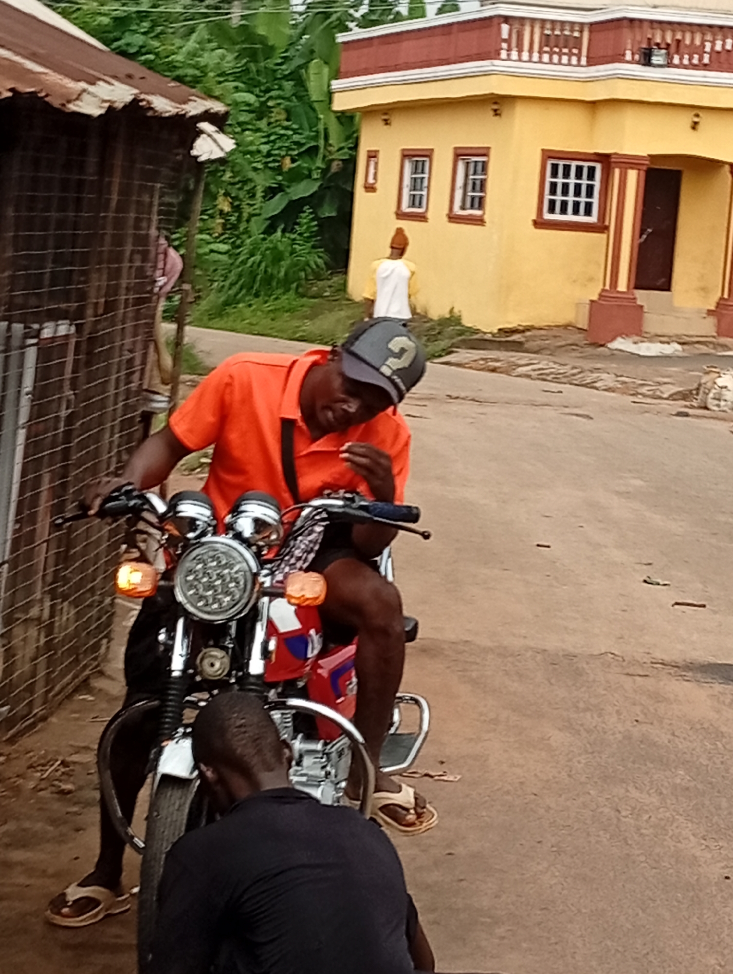 A full-shot image depicts two males on a dirt road in Nsugpe, Nigeria. In the foreground, a male wearing a black t-shirt and black pants is partially visible, bent over the front tire of a red and silver motorcycle. Behind him, the motorcycle is occupied by a male wearing an orange t-shirt, dark shorts, and a grey cap with a question mark logo. He is holding a small object in his right hand and appears to be looking down at it. The motorcycle has a large, round headlight with multiple LED bulbs, and orange turn signals. To the left of the motorcycle, a wooden structure with a corrugated metal roof and a wire fence is visible. In the background, a yellow building with red trim and a red door stands prominently. Lush green vegetation, including banana leaves, surrounds the building. The sky is overcast. The scene appears to be during the daytime.