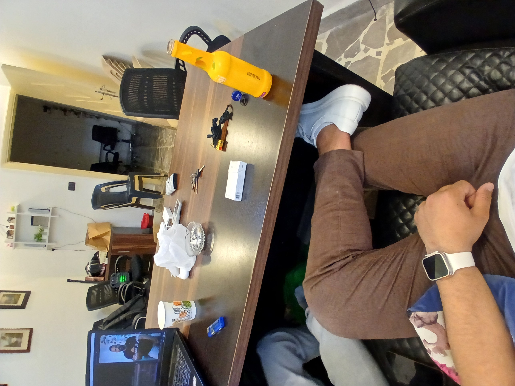A person is seated at a wooden desk indoors. On the desk are a laptop, a yellow bottle, keys, an ashtray, a cup, and a box. The person is wearing brown pants, a white shoe, and a white watch. The location is likely indoors but there is insufficient information to determine that the location is Rukn ad-Din Municipality, Syria.