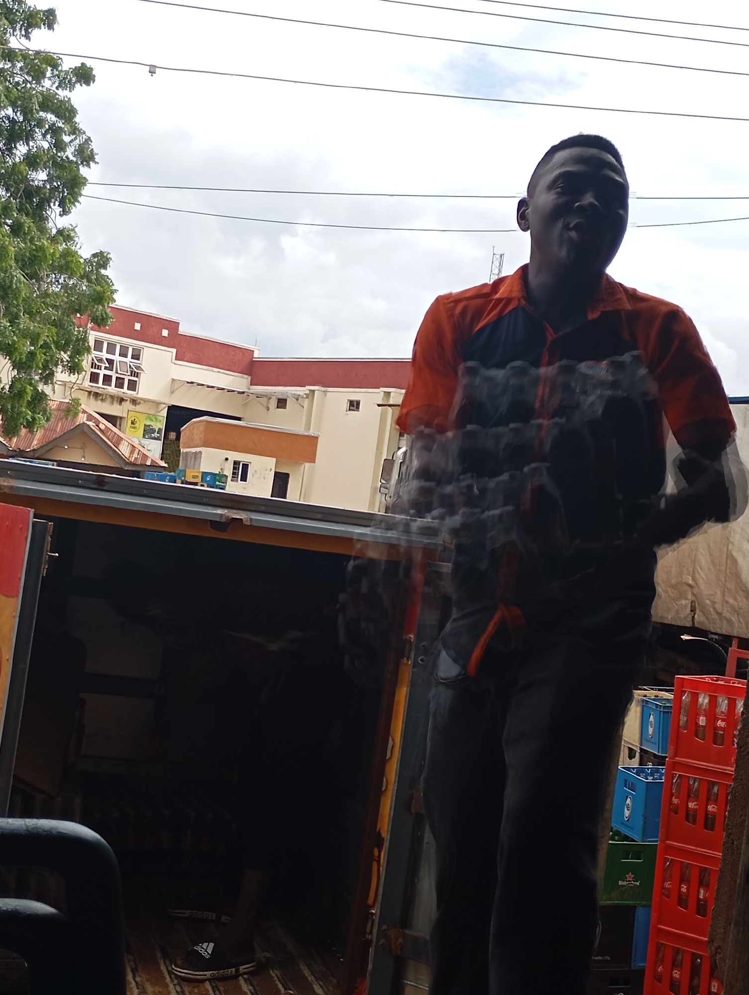 In Shendam, Nigeria, a man wearing an orange and black collared shirt and dark pants stands in front of a delivery truck filled with cases of drinks. He appears to be in motion, with his body slightly blurred. Behind the truck, a building with a red roof and cream-colored walls is visible, with several windows. The sky above is overcast. The truck bed is filled with various crates, including red ones containing bottles of Coca-Cola, blue crates with logos, and green crates. The man is standing near the open rear of the truck.