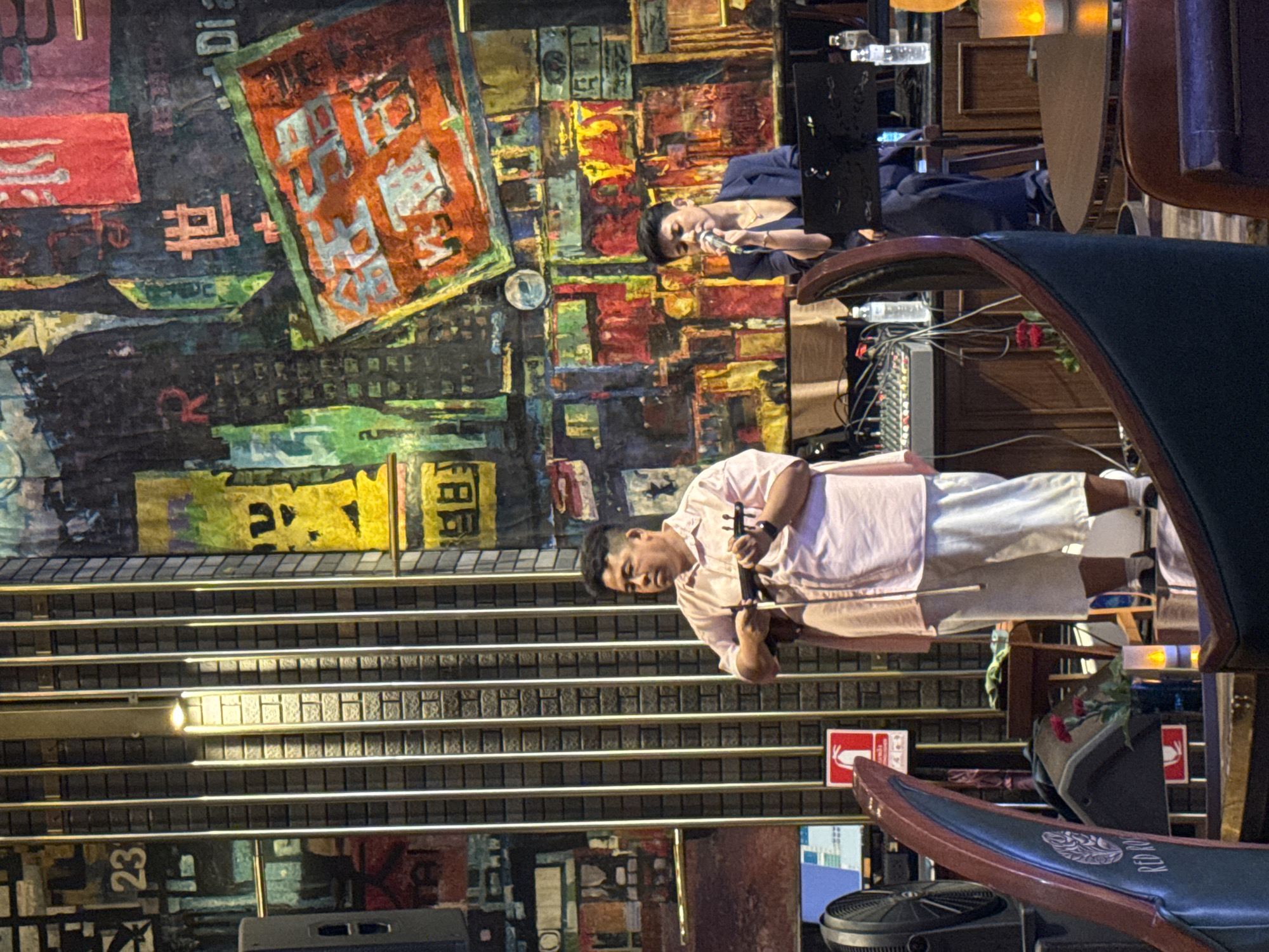 The image depicts two men in what appears to be a performance setting within a venue likely located in Bangkok, Thailand. A man in a light pink shirt and white shorts is playing a violin. Behind him, another man wearing a dark blue outfit is speaking into a microphone. Both are situated in front of a vividly colored and layered abstract mural with Asian script. The venue features a dark metal horizontal grid structure, as well as upholstered chairs and seating with a logo of a flower and "RED ROSE" visible. A sound mixer and other equipment are located nearby. A fire safety sign is also visible.