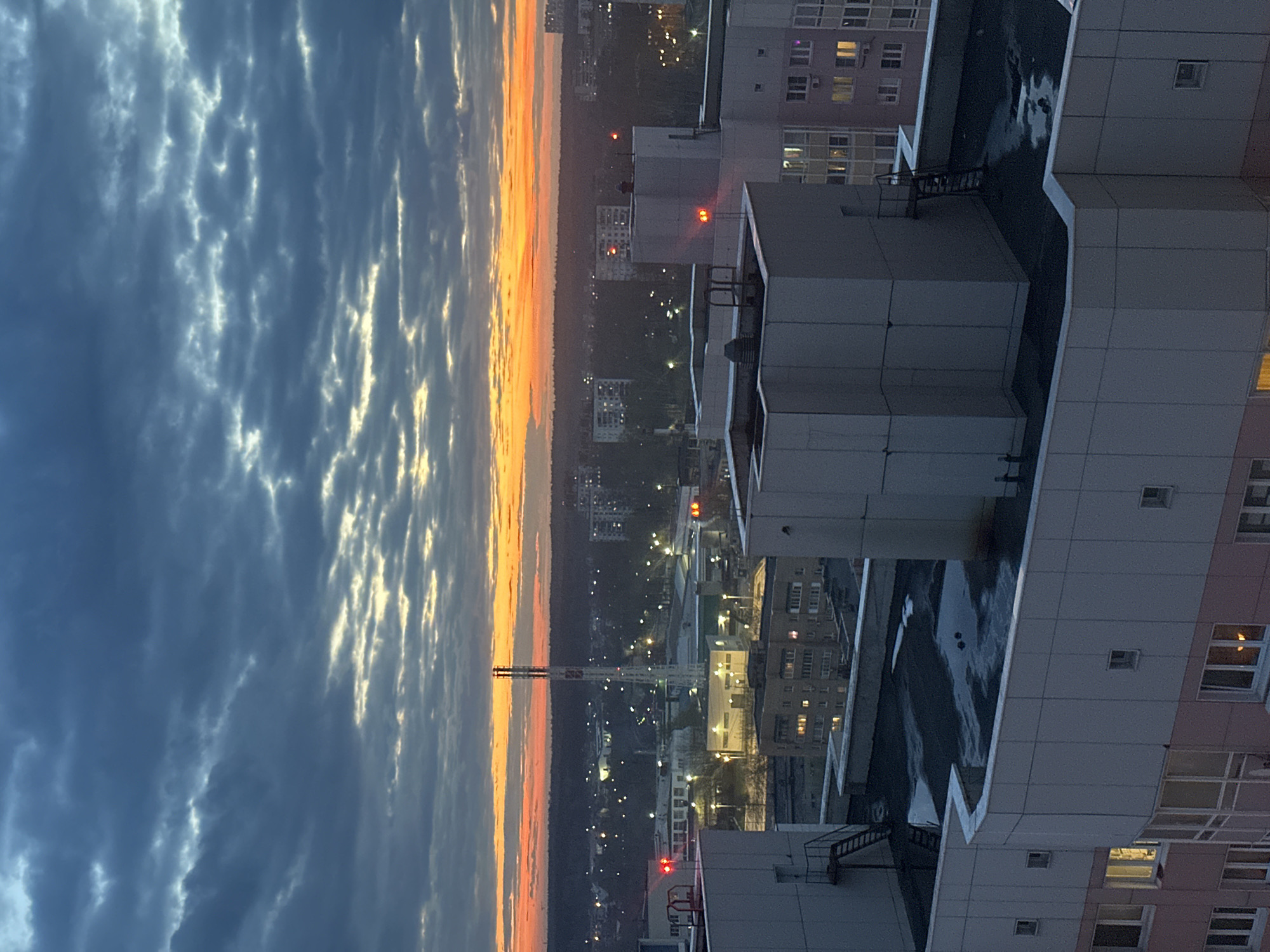 The image presents a panoramic view of an urban landscape, likely Krasnogorsk, Russia, captured during either sunrise or sunset from a high vantage point, possibly a rooftop or a tall building. The sky dominates the upper portion of the frame, filled with textured, streaky clouds in shades of deep blue and grey. A brilliant band of orange and yellow light stretches across the horizon, illuminating the undersides of some clouds and indicating the presence of the sun just below the visible line.
In the midground, several modern multi-story buildings, appearing to be residential or commercial blocks, are prominent. These buildings feature flat roofs, some of which display mechanical units or structures. Red lights, likely aircraft warning beacons, are visible on the rooftops of some buildings. Windows in these structures are scattered, with many emitting a warm glow, suggesting internal activity or artificial lighting against the dimming or brightening sky. Details like utility ladders or stairs are visible on the exterior of some buildings.
Beyond these immediate structures, the cityscape extends into the distance, marked by a multitude of twinkling artificial lights from various buildings and streets, which illuminate the urban fabric against the darker background. A tall, slender white structure, possibly an industrial chimney or communication tower with multiple flues, stands out in the mid-distance, silhouetted against the vibrant horizon. A dark band of trees or forested land is visible along the horizon line, separating the city from the colorful sky.
No explicit human activity is discernible, lending a serene and expansive feel to the urban scene. The dramatic interplay of light and shadow, coupled with the vivid sky colors, creates a striking and atmospheric view. There is no visible text in the image.