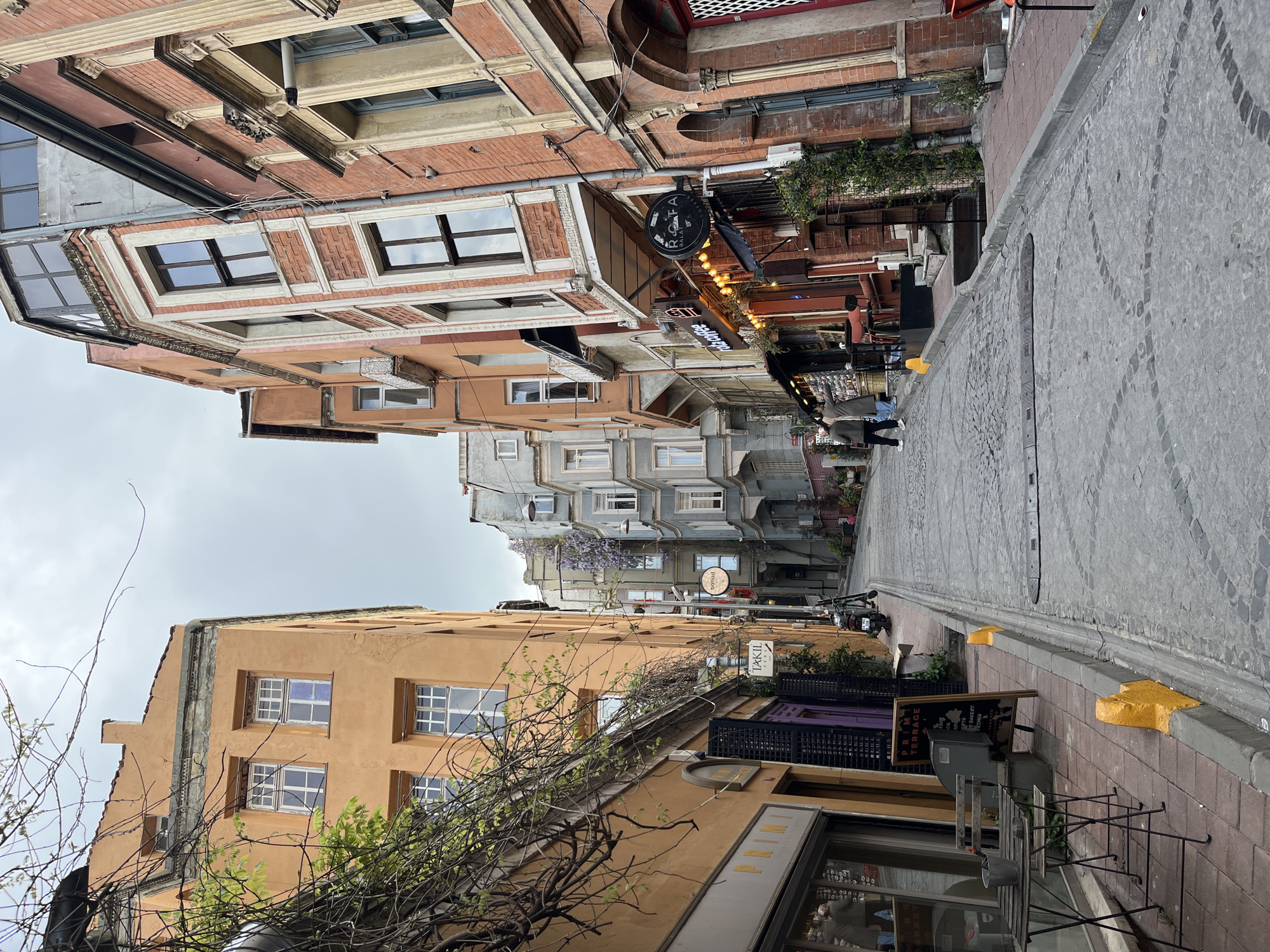 The image displays a vibrant street scene in Istanbul, Turkey, featuring a narrow cobblestone street flanked by old, multi-story buildings under an overcast sky. The street, paved with grey cobblestones, exhibits decorative patterns and appears to gently slope and curve into the distance. Sidewalks line both sides of the street.
On the left, a building with an orange-painted facade is prominent in the foreground, featuring multiple windows with white frames. Bare tree branches and some green foliage climb along its exterior. Further along this side, buildings with greyish facades are visible, one adorned with purple flowering vines, possibly wisteria or jacaranda. A vertical sign reads "TAKI L", and a smaller round sign further down the street says "baliad".
On the right side, the buildings showcase diverse architectural styles, incorporating exposed brickwork and painted surfaces, often with ornate details like cornices and decorative window frames. Several commercial establishments are apparent. A large, round black sign prominently displays "RAFA BALIK". Below it, another sign features "OLD COFFEE" or "GOLD COFFEE", illuminated by string lights. Outdoor seating, including tables and chairs, is arranged on the sidewalk. A chalkboard sign announces "PRIMI TERRACE" and lists offerings like "COFFEE..." and "BAKERY".
A few individuals are seen walking along the right sidewalk, indicating ongoing daily activity. Yellow concrete blocks are spaced along the curb on the right. The soft, even lighting suggests either an overcast day, early morning, or late afternoon. The scene evokes a sense of history and local life within a charming urban setting.