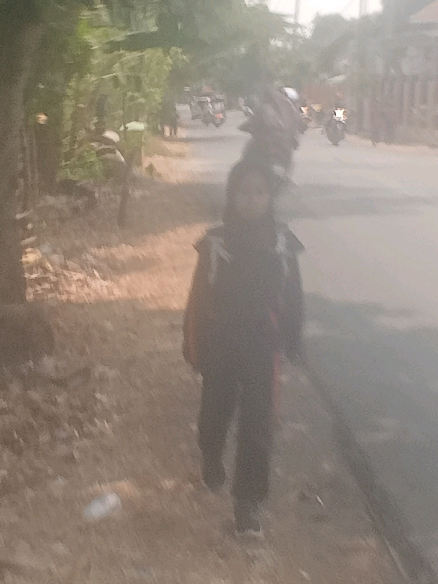 A person, possibly a child, walks along a dusty road in the city of Lebakgowah, Indonesia. The person is wearing a dark jacket and dark pants. The road is lined with trees and buildings. In the background, several motorcycles are visible on the road. The lighting suggests it is daytime, with sunlight filtering through the trees, creating a somewhat hazy atmosphere. The ground is dry and appears to be made of dirt or gravel, with some scattered debris.