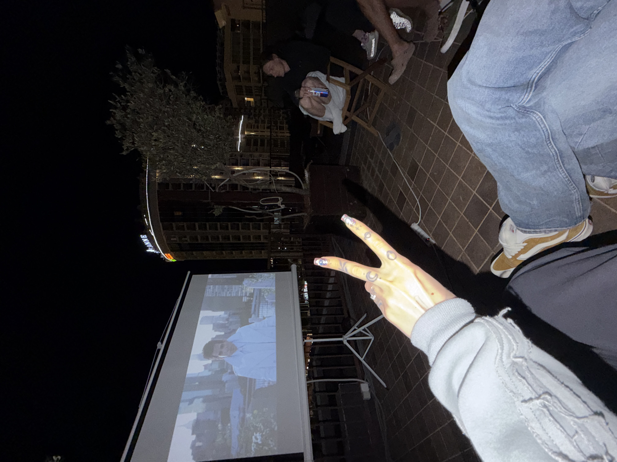 The image shows an outdoor movie night scene, likely on a rooftop in Budva, Montenegro, as indicated by the "AVAVE" sign on the adjacent building. A group of people are seated around a projected movie on a white screen. One person is making a peace sign toward the camera. The setting is at night, and the scene is lit by the projector and ambient city lights. The ground is tiled. The foreground contains the lower half of one person's blue jeans and sneakers, and another person's legs in shorts and sneakers seated in a folding chair.