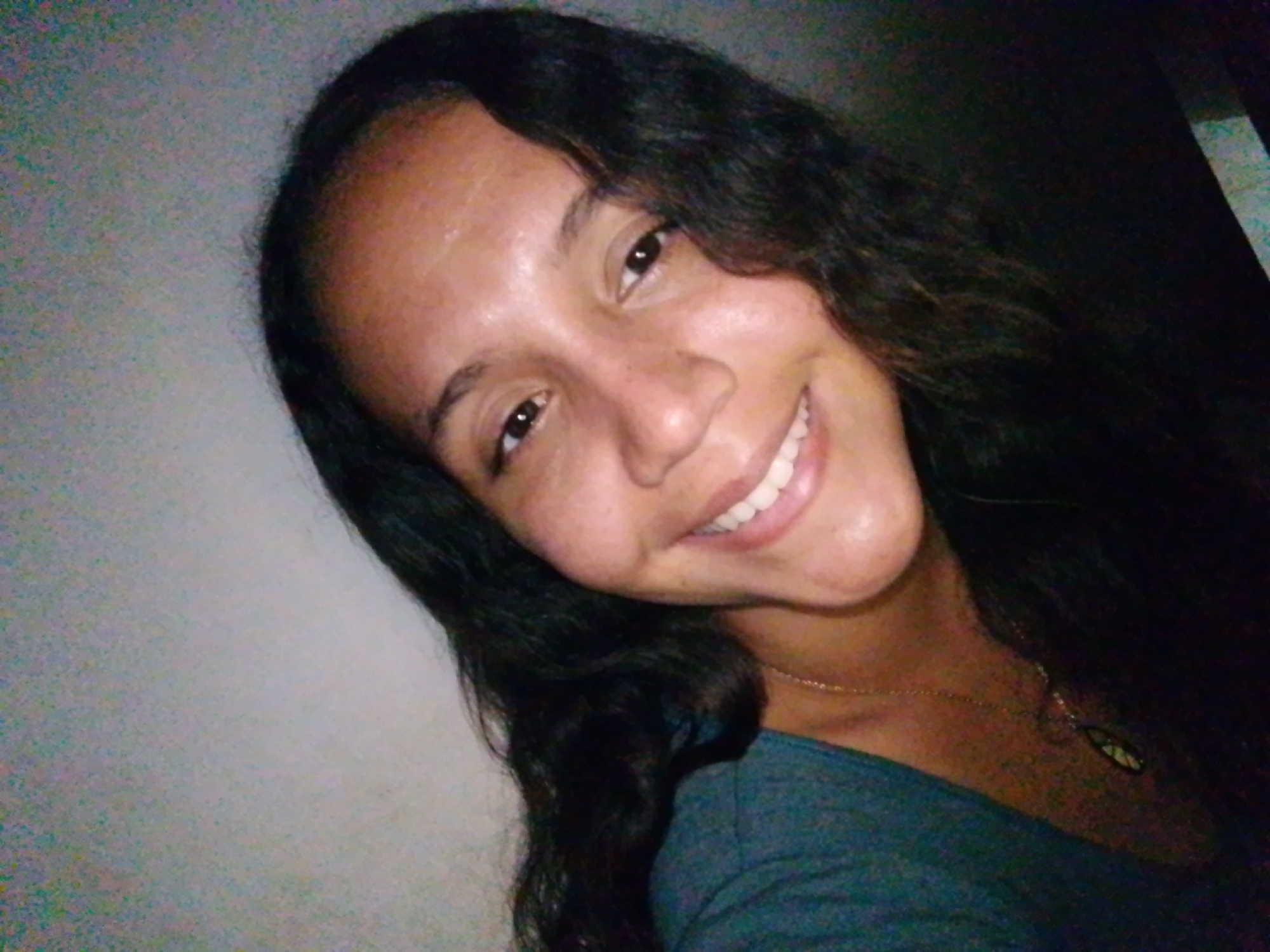 A close-up photograph shows a smiling woman with long, dark, curly hair. She is wearing a green top and a gold necklace with a pendant. The background is a light-colored wall on the left and a dark area on the right. The location, Juan Griego, Venezuela, is not discernible from the image.
