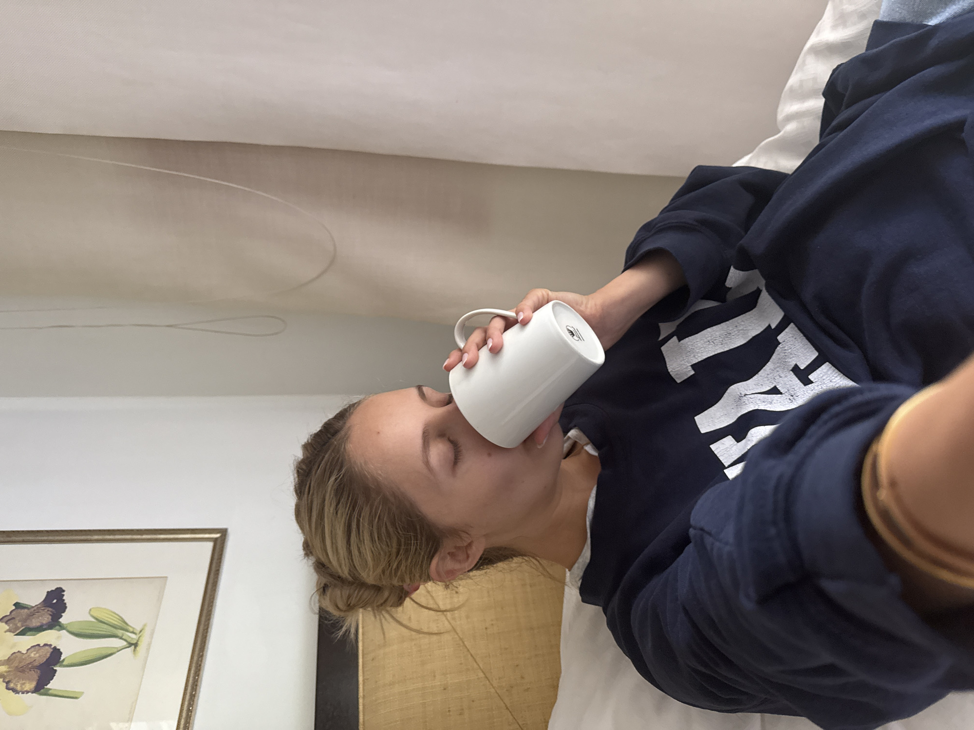A blonde woman with her eyes closed is drinking from a white mug while lying in bed. She is wearing a navy blue sweatshirt with white lettering. A framed artwork of flowers hangs on the wall behind her. The scene appears to be in a bedroom in Mexico City, Mexico.