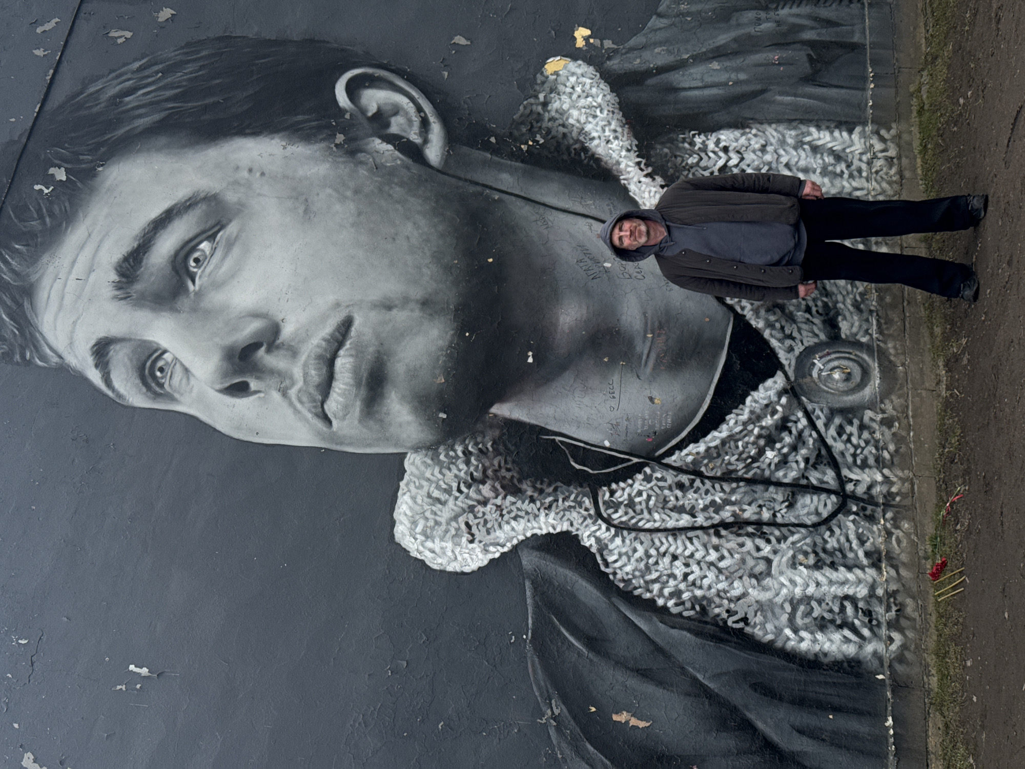 A man stands to the right of a large black and white mural on a wall in Saint Petersburg, Russia. The mural depicts a close-up of a young man's face with a knit collar. The man standing in front of the mural is facing the camera and is wearing a grey hooded sweatshirt and a black jacket and pants. His feet point to the right edge of the frame, and he is positioned between the mural and a path. A few sticks with red flowers are at the bottom right of the mural next to the path. The wall on which the mural is painted is gray, peeling, and has graffiti written in white.