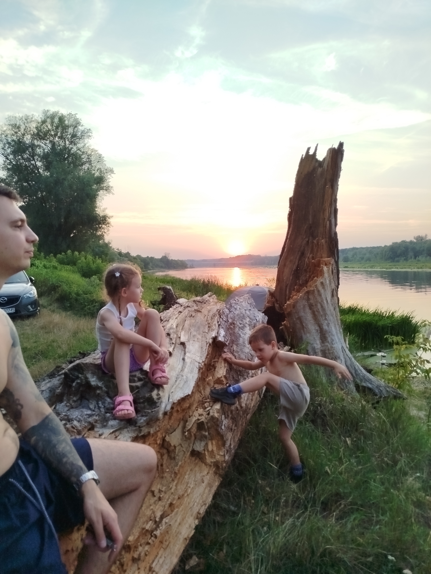 In Topolina, Poland, at sunset, a man and two children are by a river. The man, partially visible on the left, has tattoos on his arm. A young girl with blonde pigtails sits on a large fallen log. A boy is actively climbing on the log. The river flows behind them, with trees and vegetation on the far bank. A car is parked behind them on the grass. The sky is cloudy with the sun setting in the distance, casting a warm glow on the scene.