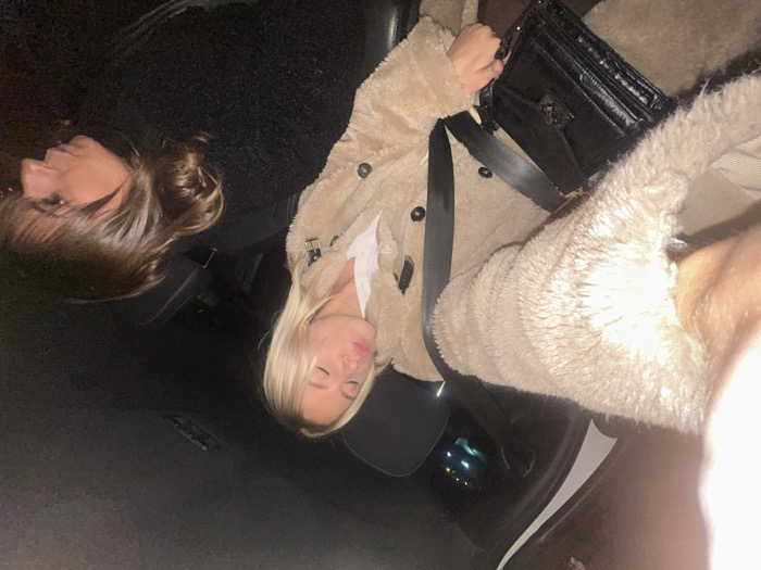 Two women are in a car. One woman has shoulder-length brown hair and is wearing a dark, fuzzy coat. The other woman has long blonde hair and is wearing a light brown, teddy bear style coat. She is also wearing a black purse across her chest. They appear to be posing for a photo inside the vehicle.