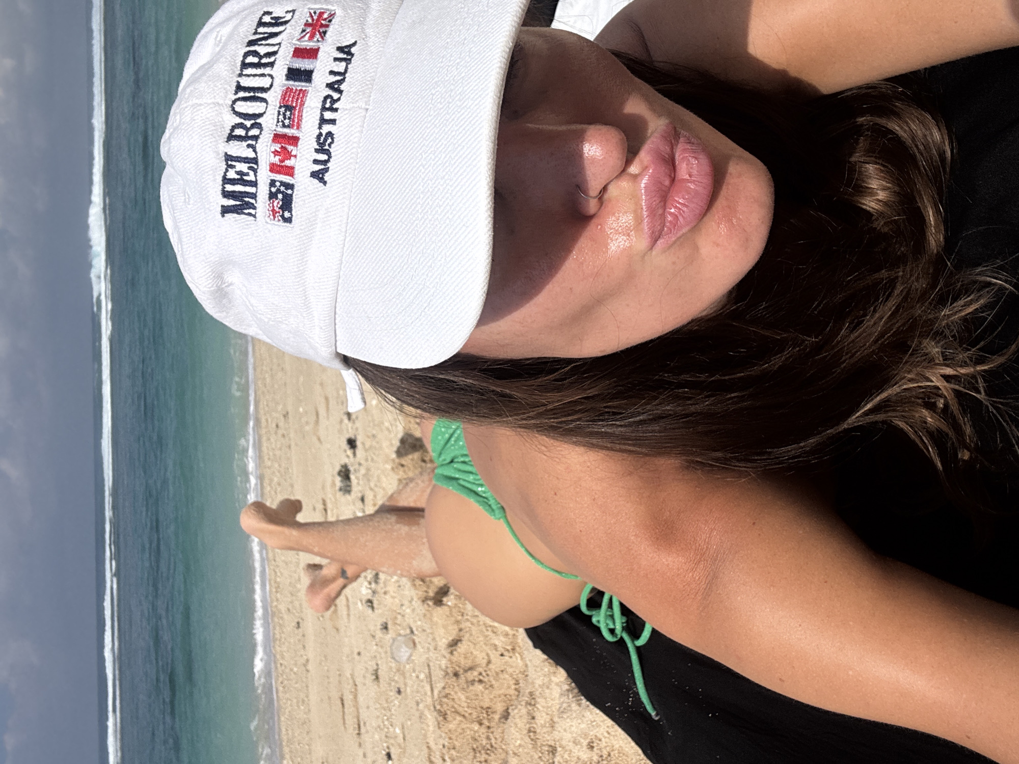 In Kuta Selatan, Indonesia, a woman with dark hair lies on a black towel on a sandy beach, facing the ocean. She wears a white baseball cap with "MELBOURNE AUSTRALIA" embroidered on it, along with several flags. She is also wearing a green bikini. The ocean is blue with white waves, and the sky is cloudy. Her legs are crossed and propped up on the sand behind her. She is looking towards the camera and puckering her lips.