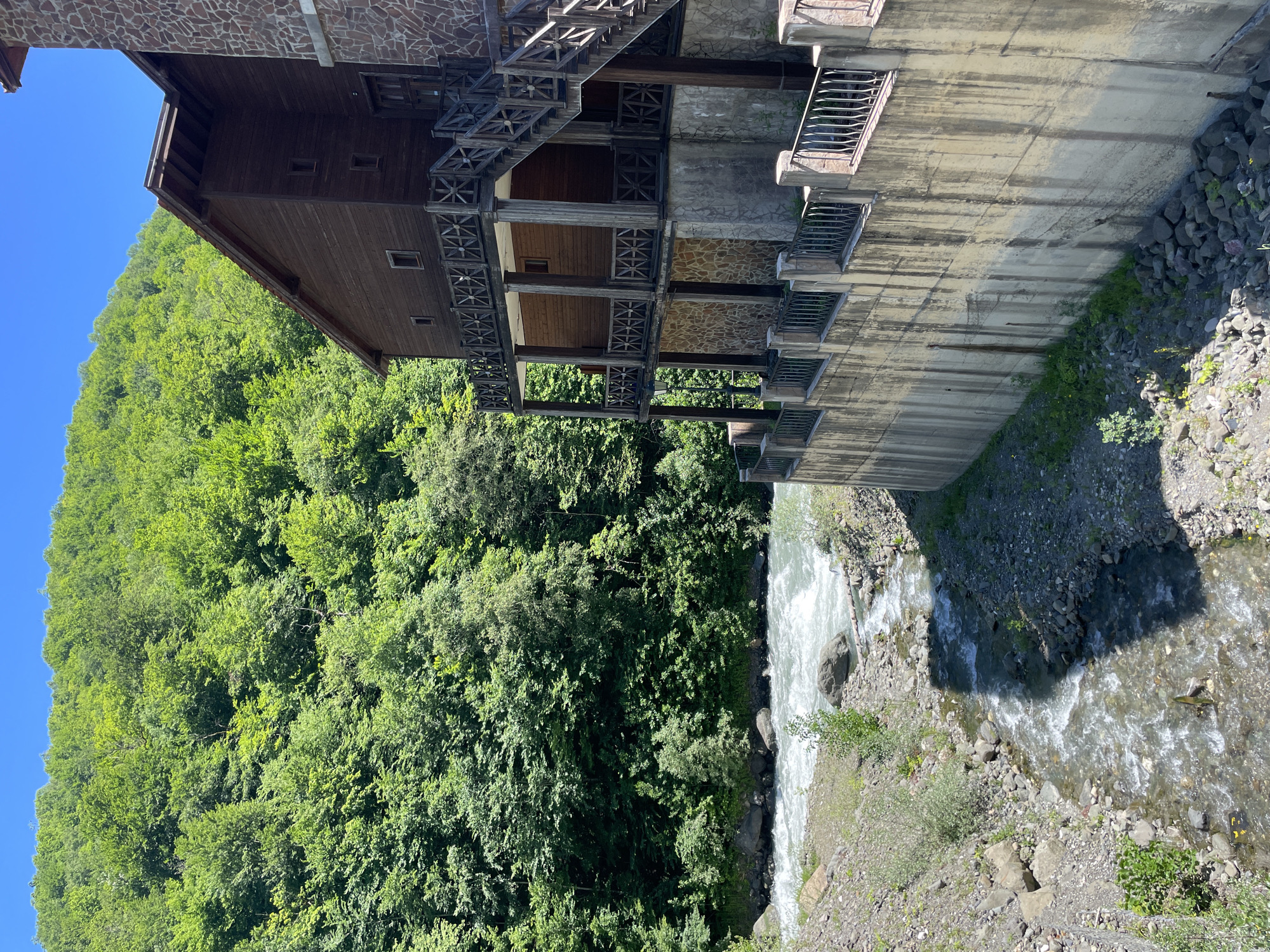 The image depicts a multi-story wooden and stone building situated beside a fast-flowing river. The building has wooden balconies with decorative railings and an external wooden staircase. The surrounding environment is densely forested, with lush green trees covering a steep hillside. The river flows over rocks, creating whitewater. The foreground shows a rocky riverbank with smaller streams feeding into the main river. A concrete wall with a decorative metal railing separates the building from the riverbank. The sky is clear and blue. The location is identified as Estosadok, Russia. There are no people visible in the image.