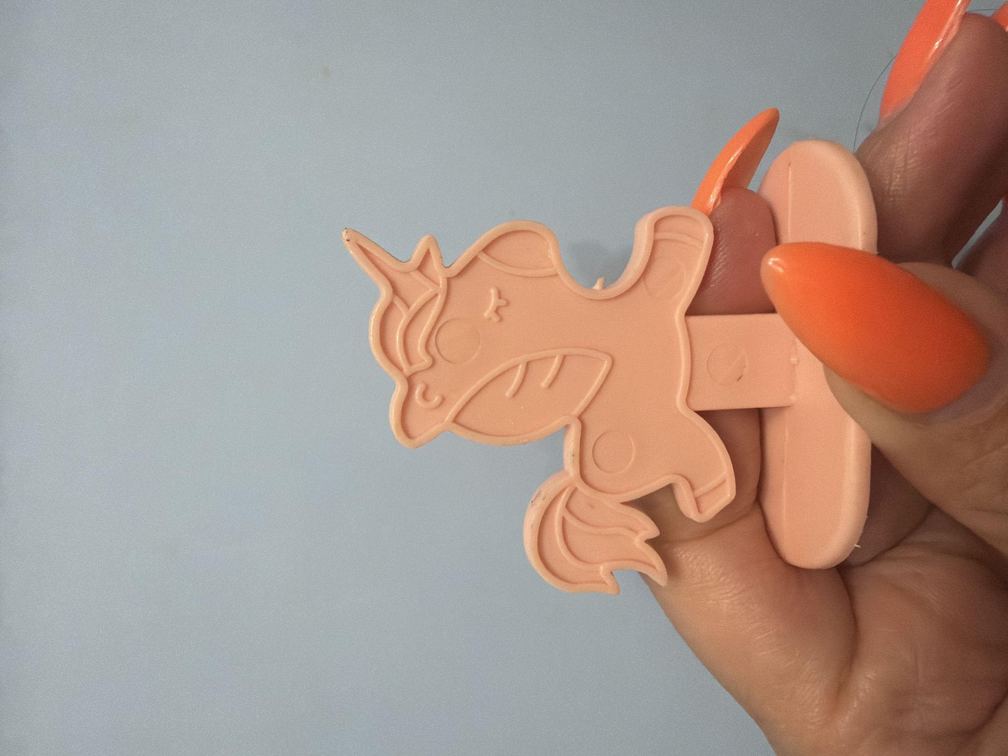 A person is holding a light pink plastic unicorn figurine with a base. The unicorn is stylized with a mane, tail, and horn. The figurine is held between the thumb and index finger of a left hand. The person has bright orange, elongated fingernails. The background is a solid light blue. The image does not contain any information about the city of Sochi, Russia.
