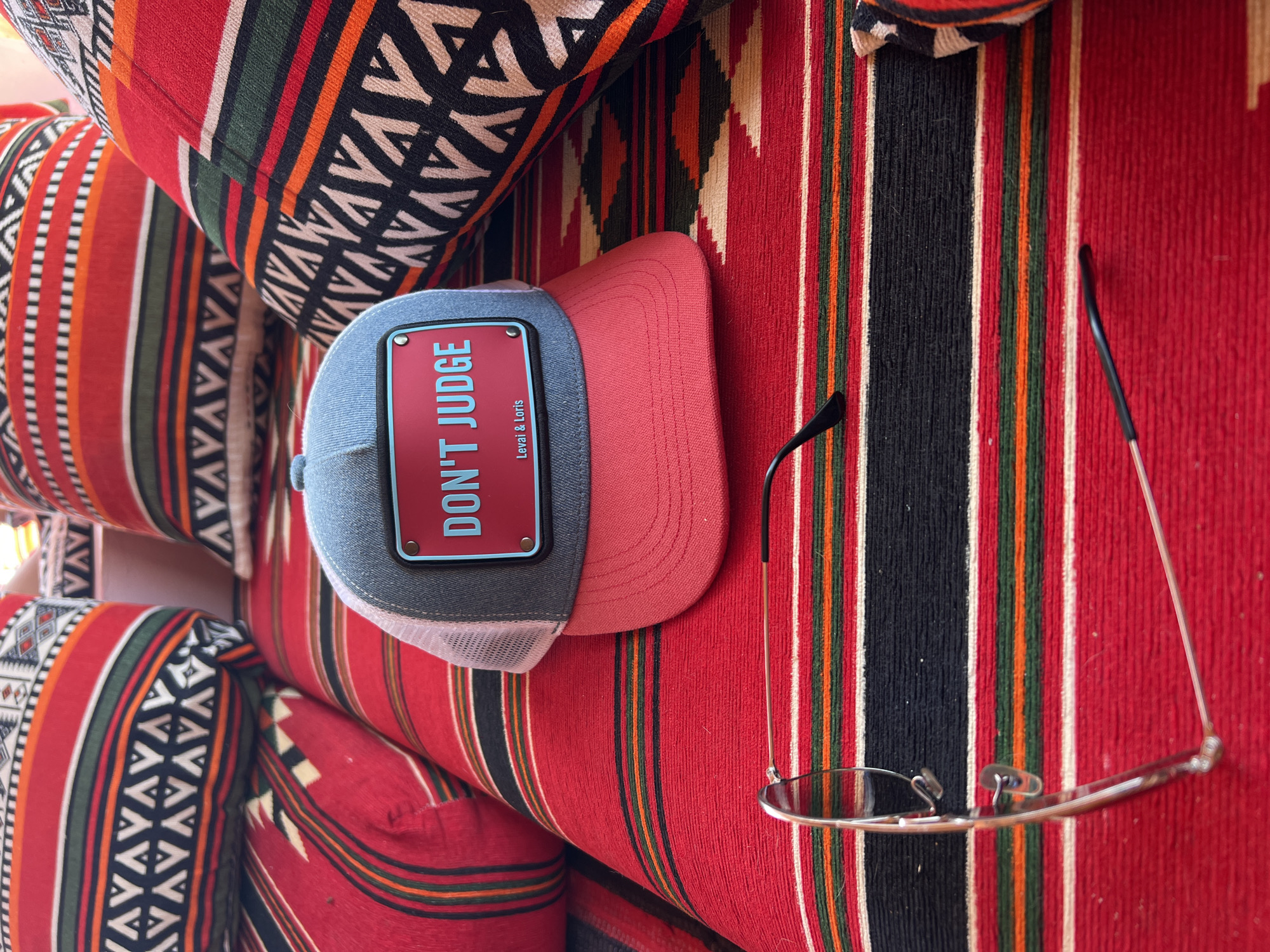The image displays a close-up, overhead view of a red and black striped fabric, likely a rug or upholstery, in an outdoor setting. A gray and pink trucker hat with a red patch reading "DON'T JUDGE Levai & Loris" sits atop the fabric. A pair of eyeglasses, with silver wire frames and black earpieces, is also present on the fabric. The scene is illuminated by natural sunlight, creating soft shadows. The background features out-of-focus pillows with similar red and black striped patterns, suggesting a casual seating area. There are no people or animals visible in the image. The location is identified as an unknown area in Egypt.
