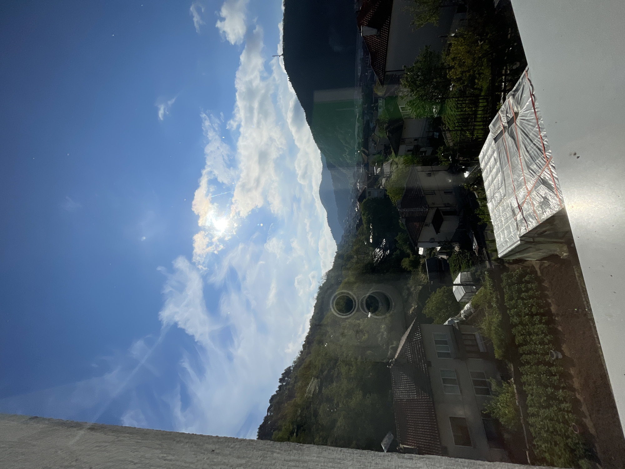 The image depicts a view through a window, showing a landscape with elements of both nature and a residential area. A mountain range is visible, partially obscured by clouds. A bright, sunny sky with scattered clouds dominates the upper left portion of the image. Below the mountains is a town with houses with red-tiled roofs. There are trees and vegetation amongst the residential buildings. There appears to be some kind of covered object or structure near the window in the bottom right. The image is rotated counterclockwise by 90 degrees. The location is in or near Zlatograd, Bulgaria, as evidenced by architectural style, mountainous terrain, and the layout of the town, as well as the image metadata indicating the location.