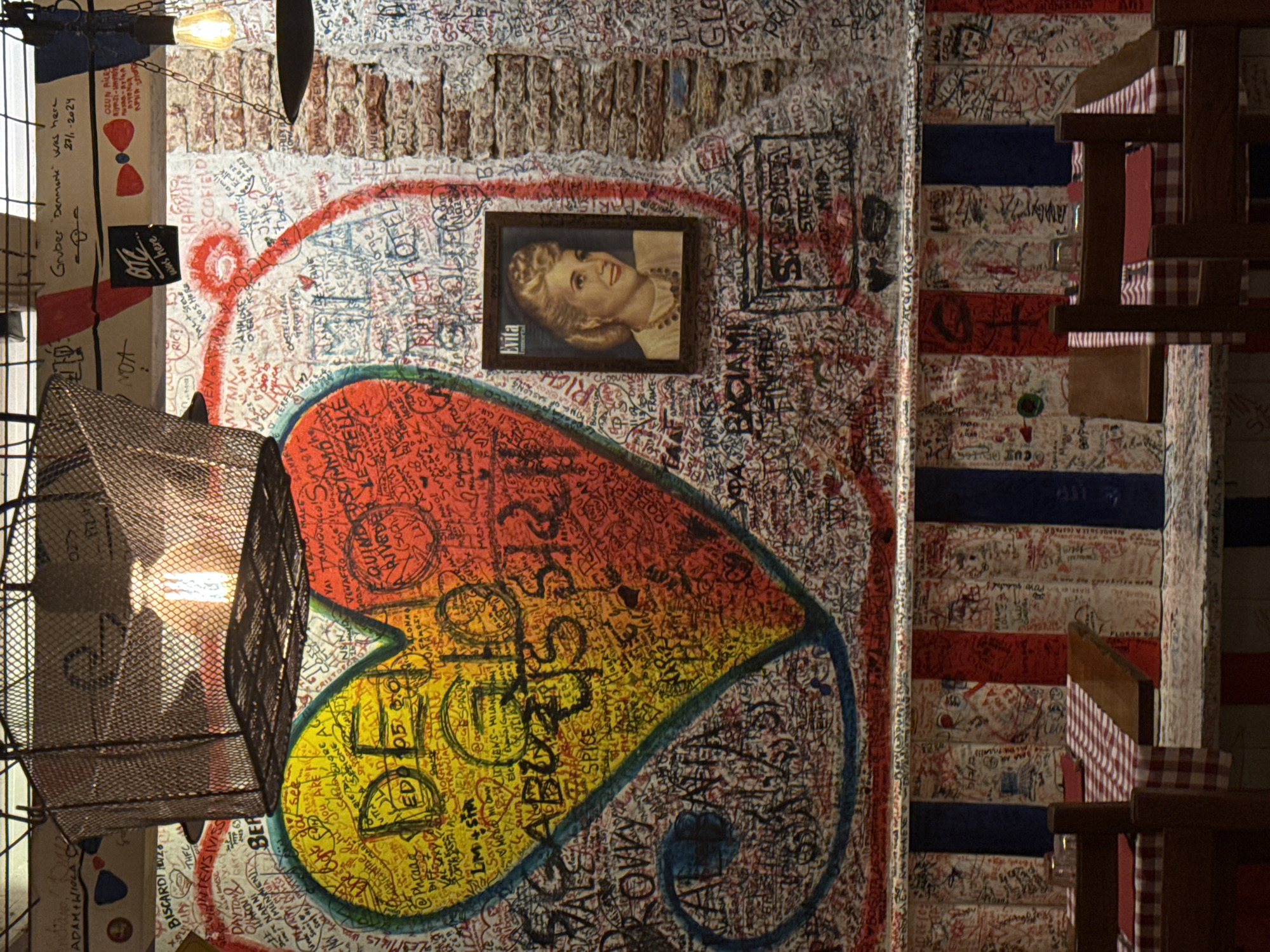 The image shows a section of a wall in what appears to be a restaurant or bar interior in Milan, Italy, due to the presence of graffiti in Italian. The wall is covered with graffiti and signatures, mostly in red ink. There is a large heart painted on the wall, split into red and yellow sections. A framed portrait of a woman labeled "Evita" hangs on the wall above the heart. A metal-caged light fixture is partially visible in the foreground, obscuring some of the artwork. The lower portion of the wall has red and blue stripes. Some wooden chairs and tables with red-and-white checkered cloths are positioned against the right edge of the frame.