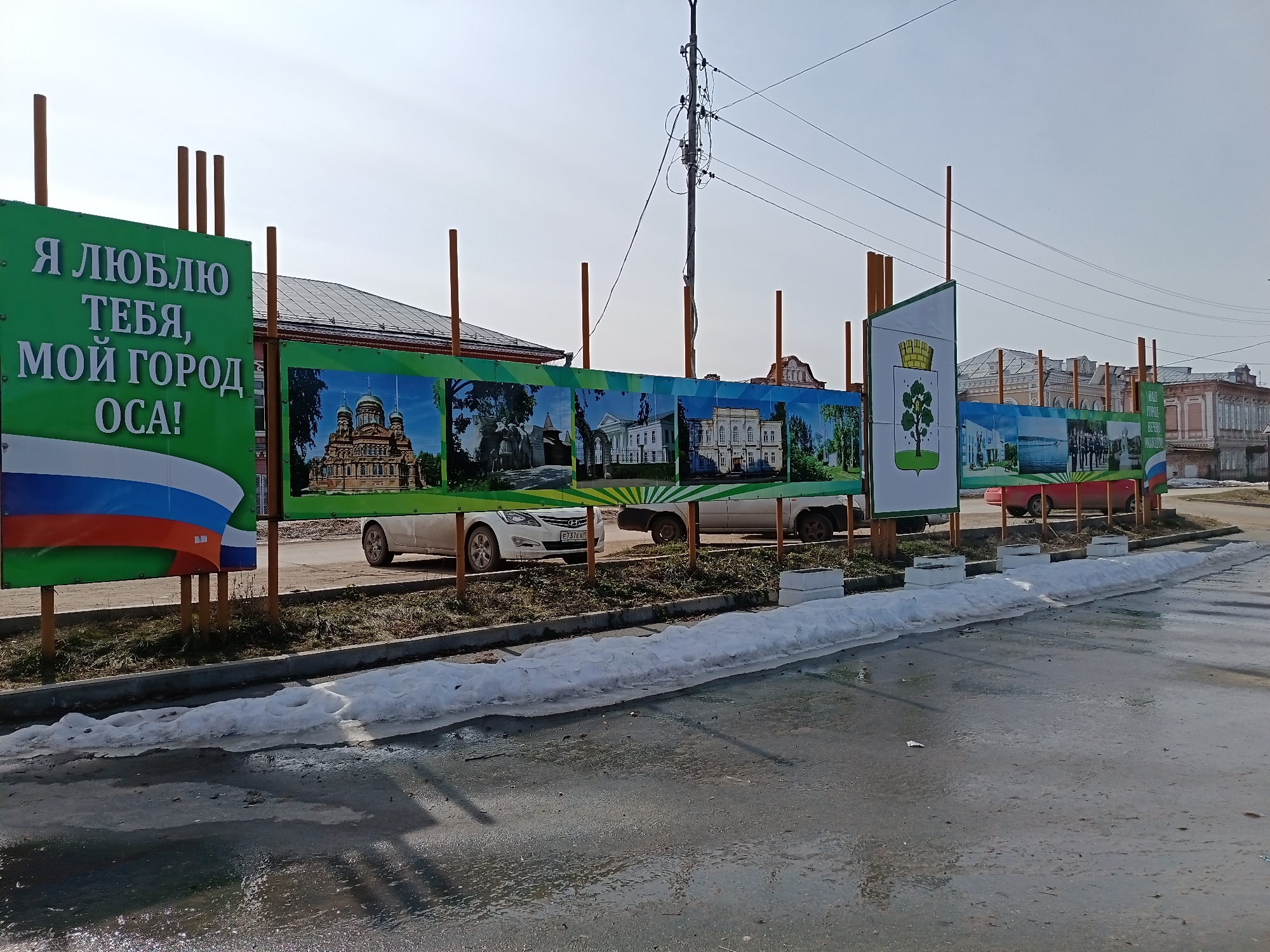 The image depicts a street in Osa, Russia, likely in early spring. Patches of snow and slushy water cover the ground. A series of green and brown billboards line the street, displaying images of local landmarks. The first billboard on the left has white lettering in Russian, "Я люблю тебя, мой город Оса!" (I love you, my city Osa!), along with a representation of the Russian flag. The billboards feature pictures of local architecture including churches and official buildings. Cars can be seen parked behind the display. A utility pole with electrical wires is visible in the background. The buildings across the street are two stories tall with brick facades.