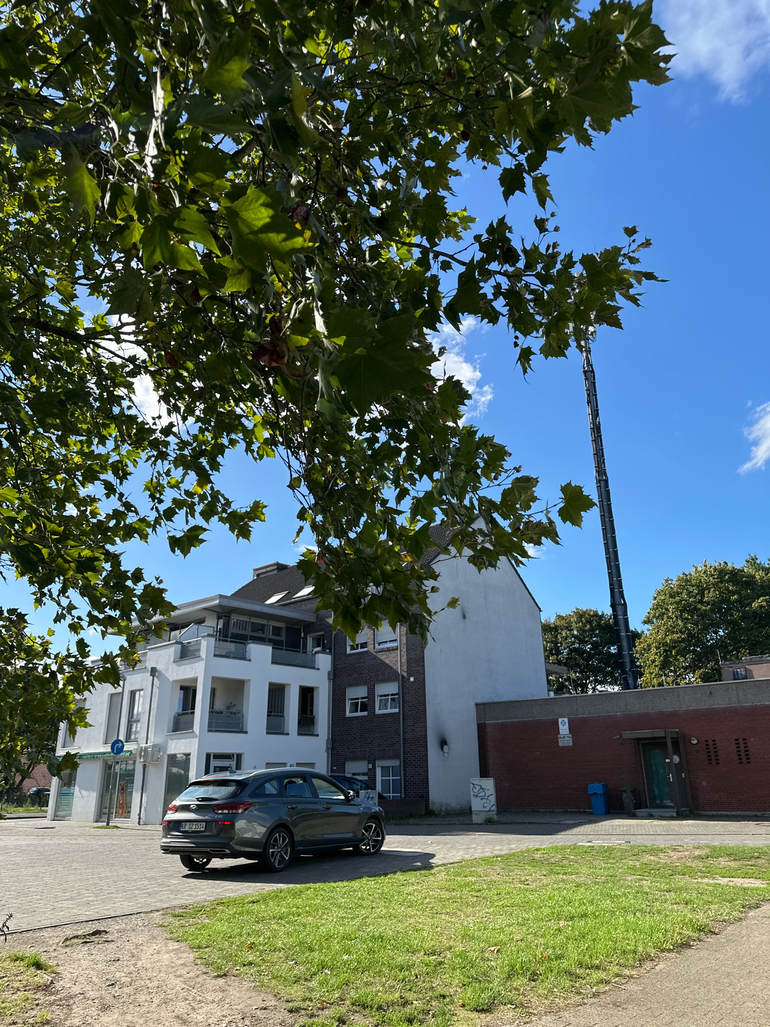 The image shows a street scene in Krefeld, Germany, on a clear day with blue skies and scattered clouds. In the foreground, a dark gray station wagon is parked on a paved area. To the left, a large tree with green leaves partially obscures the view of a modern white building with multiple balconies. Behind the white building, a brick building is visible. Further in the background, a tall, slender communications tower stands against the sky. The ground is a mix of paved surfaces and a grassy area. The scene appears to be in a residential or mixed-use urban area.