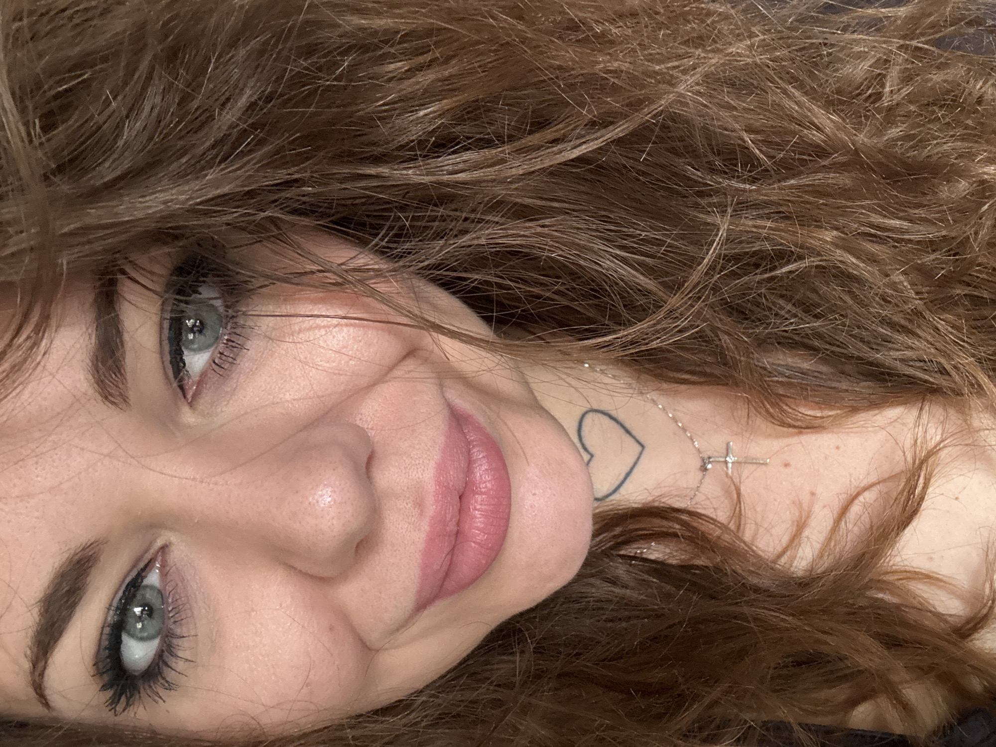 A close-up, chest-up portrait of a woman with light brown, wavy hair. Her eyes are light green, and she is wearing dark eyeliner and mascara. Her lips are painted with a light pink lipstick. She has a small heart tattoo on her neck and is wearing a silver necklace with a cross pendant. The background is a dark, blurred color. The image does not contain any information that would allow for the determination of its location in Kremenchuk, Ukraine.