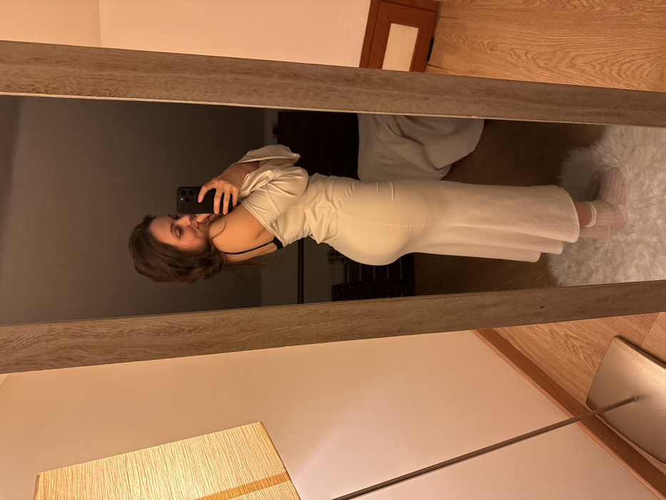 An indoor scene depicts a female individual, with medium-length dark brown hair, taking a mirror selfie. She is positioned to her right side, facing a full-length mirror with a light brown frame. She wears a light-colored, form-fitting off-the-shoulder garment that extends below the knee, revealing a black bra strap on her left shoulder. She holds a dark-colored smartphone in her left hand, pointing it towards her reflection, and her right hand touches her chin/mouth area as she displays a subtle smile. Her feet, clad in white translucent socks, rest on a white fluffy rug positioned on a light-colored wooden floor. To the immediate left of the mirror, a floor lamp with an orange rectangular fabric shade is visible. The reflection in the mirror's background shows a dimly lit room with a warm ambient light, featuring a dark furniture piece, possibly a bed or dresser, partially covered by a light-colored textile. The specific location within the city of Cava de' Tirreni, Italy, cannot be determined from the provided visual information.