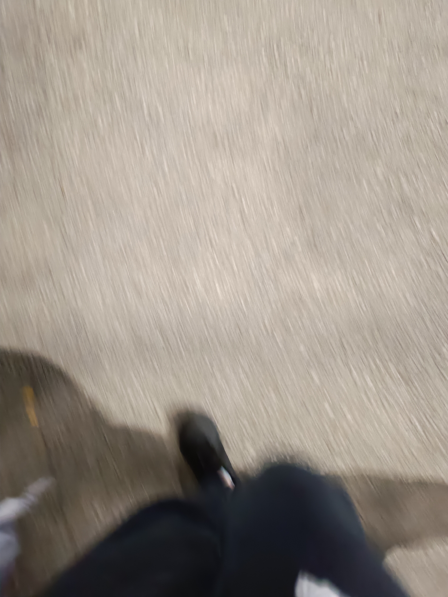 The image shows a blurred first-person perspective of a person walking on a paved surface. The person is wearing dark-colored pants and what appears to be a black shoe. The pavement is light-colored and exhibits linear texture. There is also a shadow and a white object at the edge of the frame. There is no information present in the image to deduce whether or not the photo was taken in Pleven, Bulgaria.