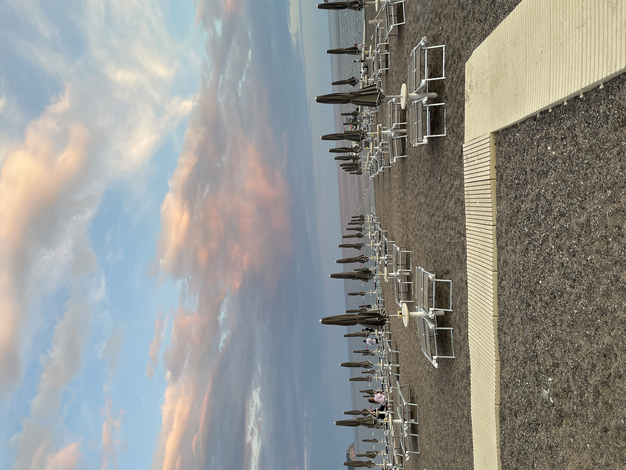 The image shows a beach with rows of white lounge chairs and dark umbrellas extending into the distance. The sand is dark in color, possibly volcanic, and a light-colored textured walkway borders the beach. The sky is filled with pink, grey, and blue clouds. The location is Rafailovići, Montenegro.