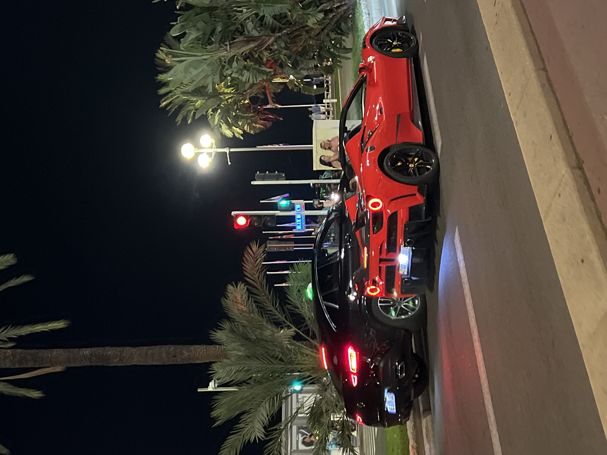 At night, on a city street in Nice, France, two luxury cars are parked side-by-side. The car on the left is a black Mercedes-Benz GLE Coupe, its brake lights illuminated. The car on the right is a red convertible Ferrari, also with its brake lights on. Both cars are facing away from the camera. The street has yellow and white lane markings. In the background, palm trees and tropical foliage are visible. Streetlights and traffic signals are also present, with the traffic light in the upper left displaying red. Several people are standing and walking on the sidewalk in the background, near the palm trees. A sign that reads "BLUE BEACH" is visible.
