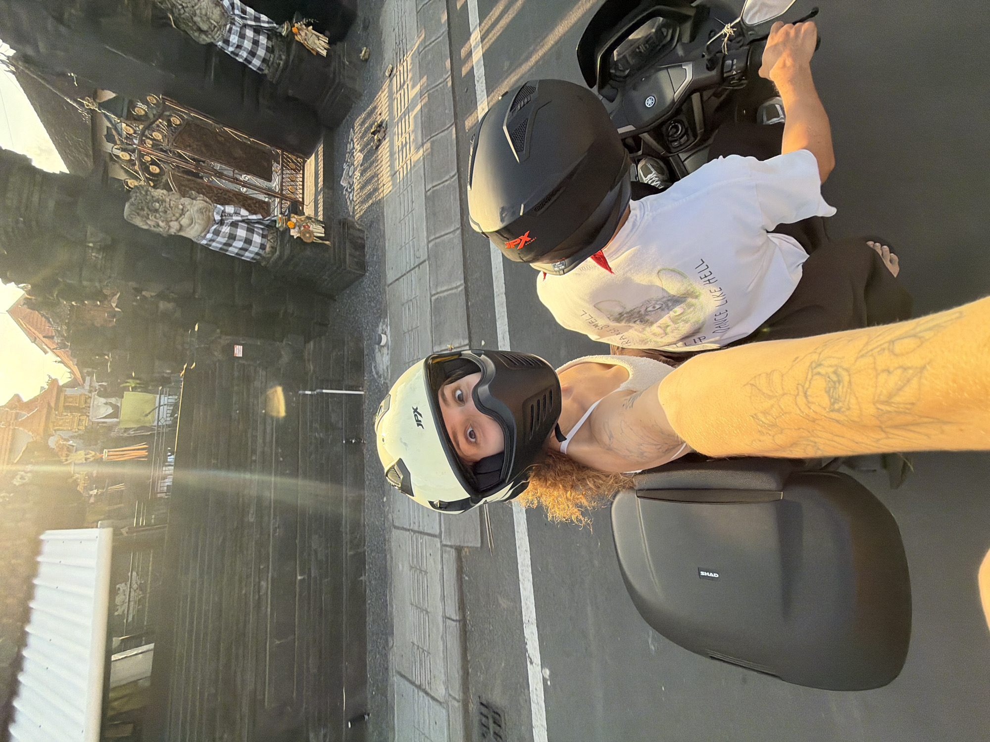 In a selfie taken from the perspective of a motorcycle rider, two individuals are captured from the chest up. The rider, wearing a white tank top and a cream-colored JPX helmet with a black face mask, faces the camera with a surprised expression. Their arm, covered in tattoos, extends forward, holding the camera. Behind them, the passenger, wearing a white t-shirt with a graphic design and a black helmet, is visible from the shoulders up. The motorcycle, a dark scooter-type, features a black storage compartment on the left. The background reveals a street scene in Mengwi, Indonesia, with a steep set of stone stairs leading up to a temple or traditional building. Ornate gates with checkered cloths draped over stone lion statues and decorative elements adorn the entrance. The setting sun casts a warm glow across the scene, creating long shadows.
