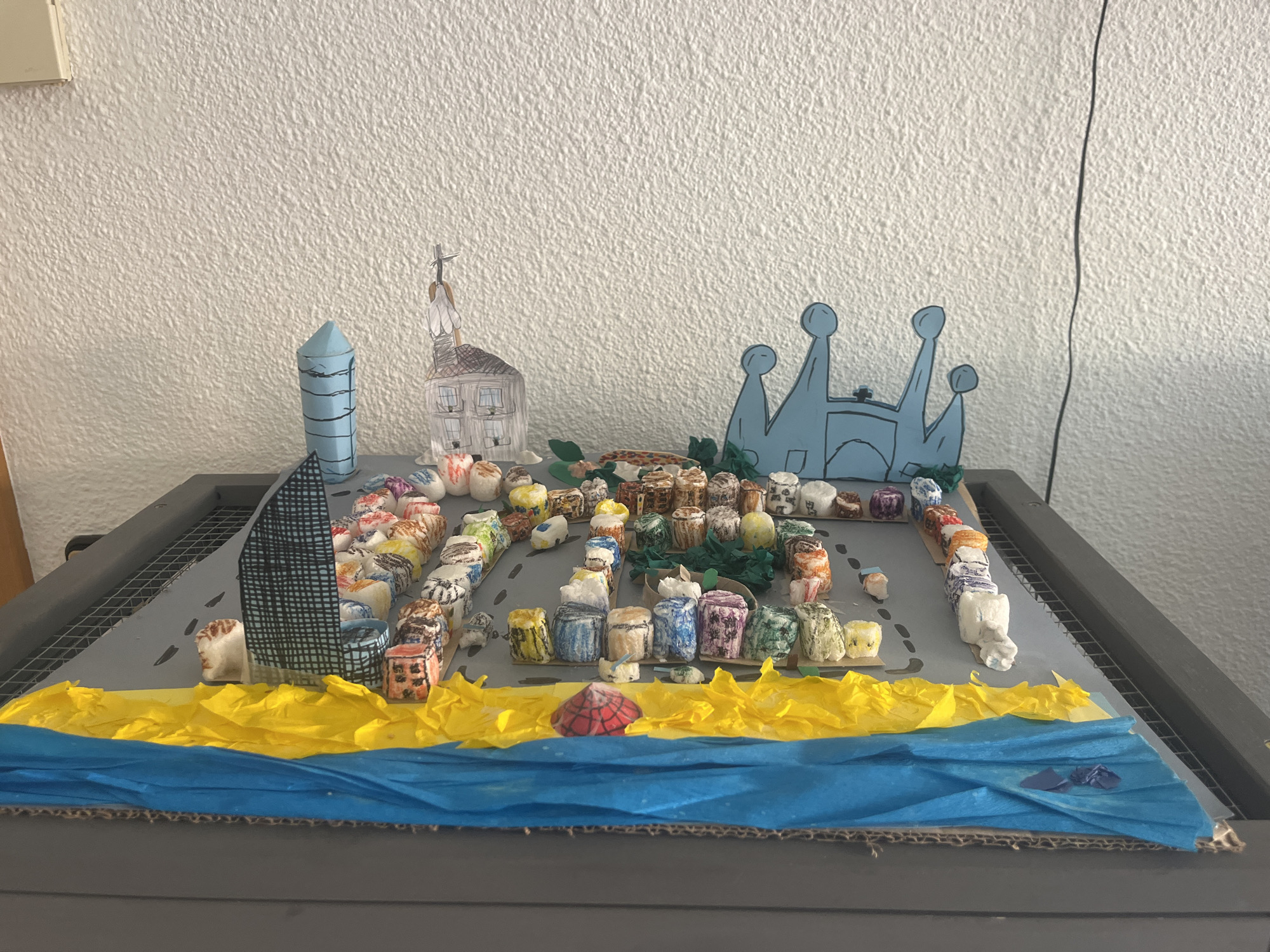 The image depicts a diorama of Barcelona, Spain. The diorama consists of a cardboard base layered with blue and yellow tissue paper to represent the Mediterranean Sea and a beach. Buildings are represented by small, cylindrical objects, painted in various colors. Some notable landmarks include a blue cylindrical representation of the Agbar Tower, a paper cutout of the Sagrada Familia, and a blue cardboard cutout of the Palau Nacional. A small, red-and-white polka-dotted parasol stands on the "beach." The diorama rests on a gray wire-framed table.