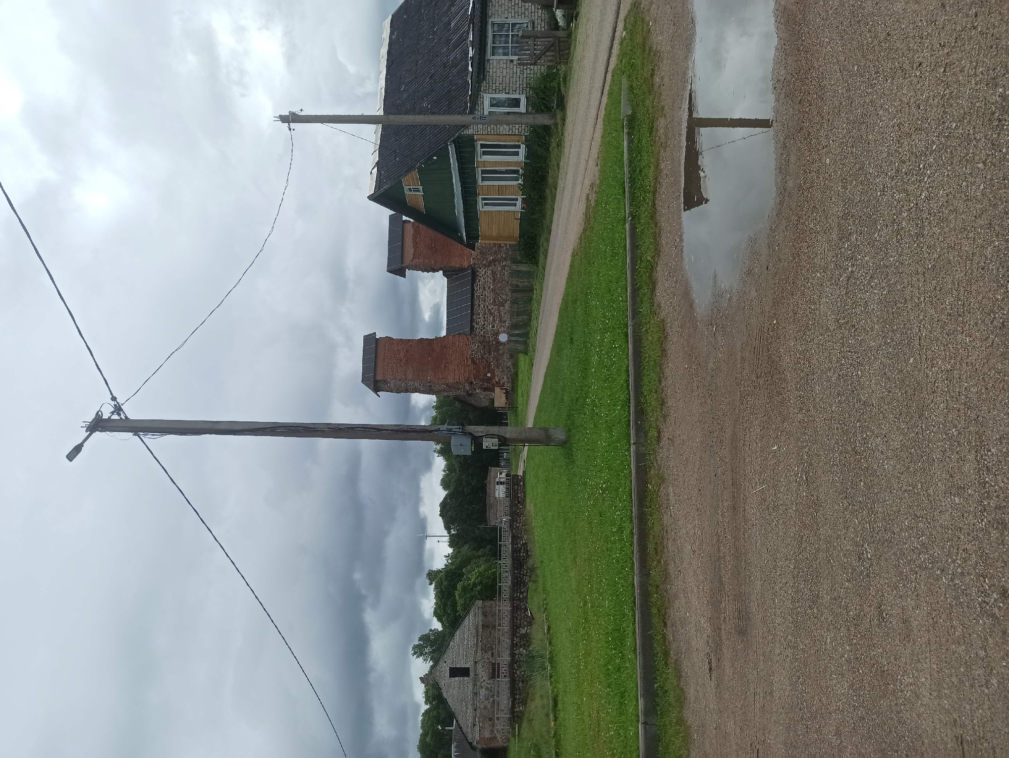 The image depicts a rural scene in the Крэўскі сельскі Савет, Belarus, under a cloudy sky. A gravel road runs through the foreground, with a large puddle reflecting the overcast sky and a utility pole. To the left of the road, a grassy area is bordered by a low stone wall and a building constructed from stone and light-colored brick. Beyond this building, trees form a backdrop. To the right of the road, a house with green and light-colored wooden siding and a dark, sloped roof stands prominently. Adjacent to the house are brick ruins, with arched openings and dark, sloped roofs. A second, smaller utility pole is visible near the house. The overall impression is one of a historic or agricultural area in a state of transition or preservation.