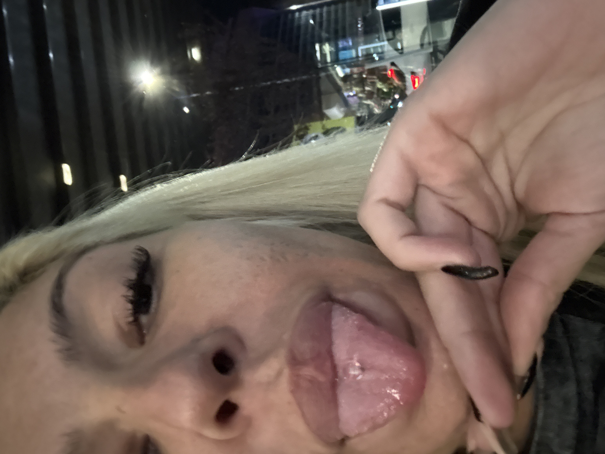 A close-up, distorted photograph depicts a light-skinned woman with blonde hair and prominent eyelashes. Her left eye is visible, closed, and she is sticking her tongue out. Part of her face, including her nose and cheek, is in view. Her hand, with nails painted black, is visible near her mouth.
The background is blurred, but it appears to be an urban environment at night. Buildings with lit windows and possible streetlights can be discerned. The city and exact location are unknown.