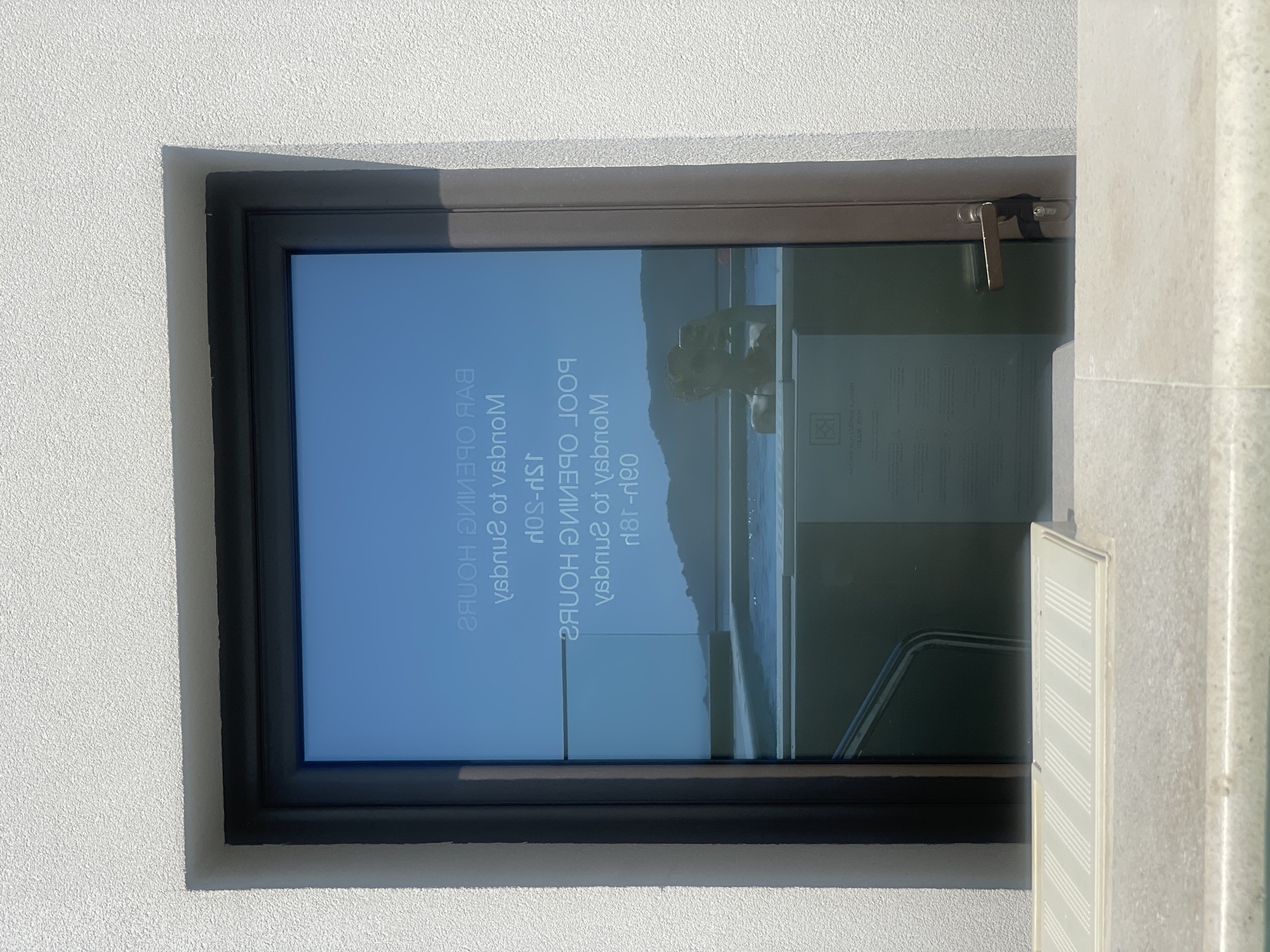 A glass door with a dark frame is set into a textured white wall. The glass is reflective and displays text in white, seemingly printed or stuck onto the glass. The text reads "BAR OPENING HOURS" and "POOL OPENING HOURS," with associated days and times that are partially obscured and reversed due to the reflection. Below the text, a reflection shows a person in sunglasses holding up a phone, taking a selfie while in a body of water, possibly a pool. In the background, there is a view of hills and a clear blue sky. In front of the door, on a ledge, is a beige object with a textured surface. The scene appears to be outdoors in a sunny location, likely a resort or hotel in Bečići, Montenegro.