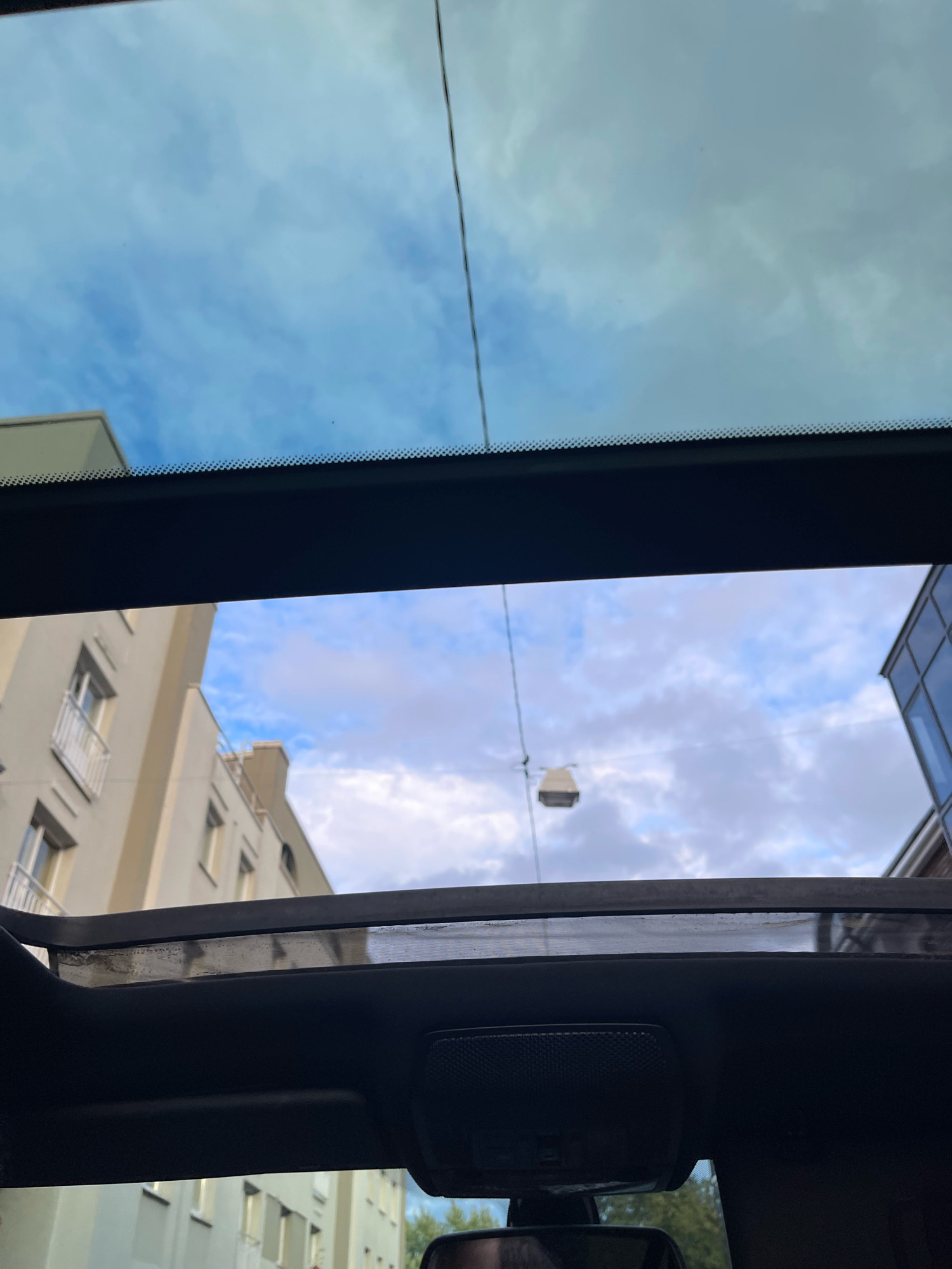 The image depicts a view through a car's sunroof, looking up at the sky and buildings. The sky is partly cloudy with a mix of blue sky and white clouds. In the distance, there are several multi-story buildings, appearing to be residential or office spaces. One building on the left has balconies with white railings. On the right, part of a modern building with large windows is visible. A street lamp hangs from wires in the middle distance. The foreground shows the interior of the car, including the sunroof mechanism and a rearview mirror. The image was taken in Tallinn, Estonia.