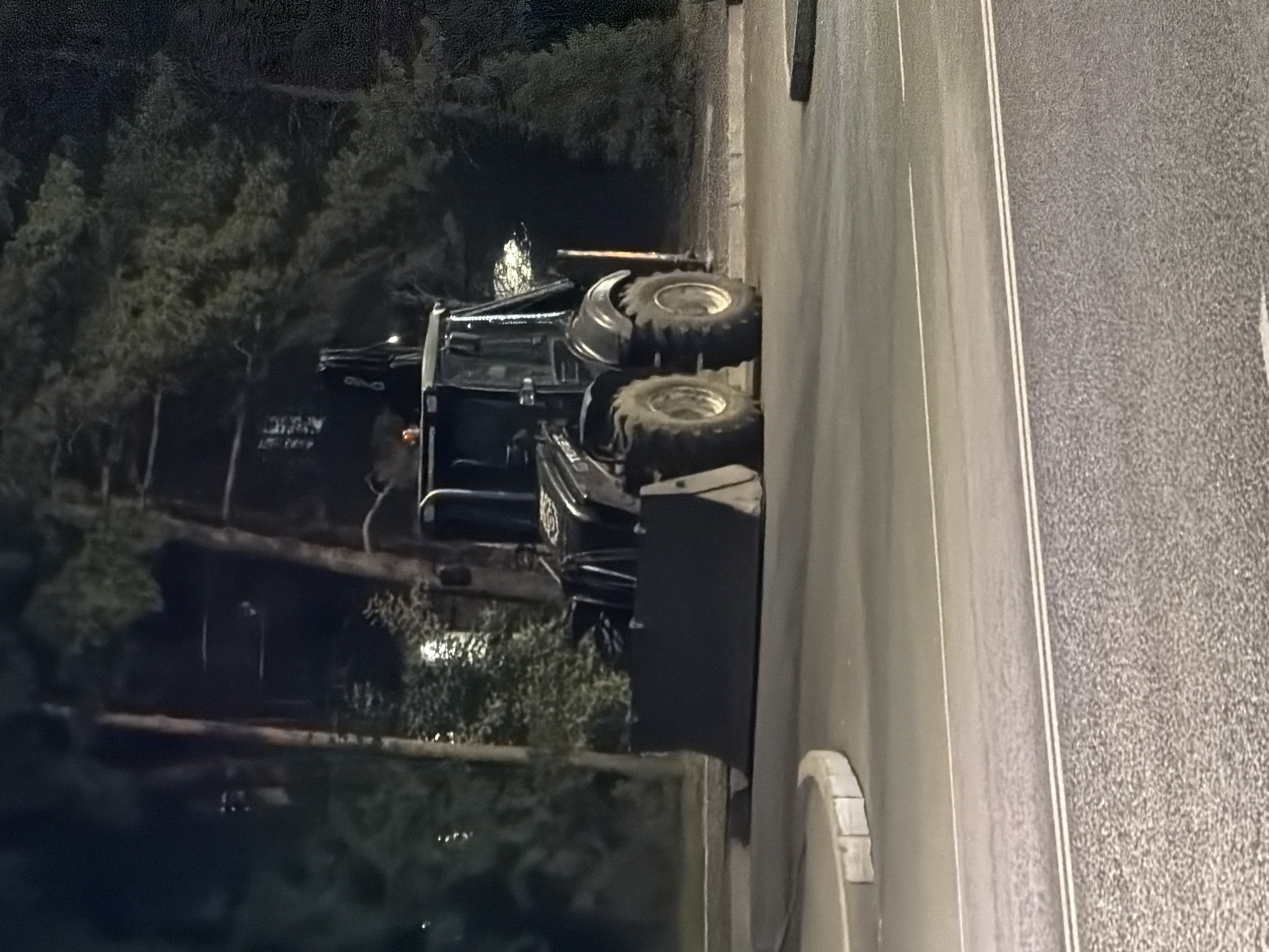 An outdoor, nighttime, medium shot shows a black backhoe loader parked on an asphalt surface, possibly a street or road, with a curb on the left. The backhoe is stationary and facing the viewer. Its front loader is lowered and resting on the ground. Behind the backhoe, trees and foliage are visible, suggesting a park or roadside setting. The scene is dimly lit, with some artificial lighting reflecting off the road surface and the backhoe. The location is identified as Pavlodar, Kazakhstan.