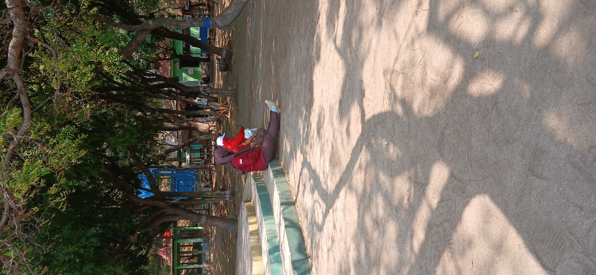 In Banyuglugur, Indonesia, two individuals are observed sitting on a tiered stone structure in an outdoor area. The person in the foreground is wearing a dark red long-sleeved shirt, dark pants, and a dark headscarf, with their legs extended forward. To their right, another individual wears a red long-sleeved shirt and a white cap, also seated. The background features numerous trees with lush green foliage, casting shadows on the sandy ground. Several small buildings with red roofs and brightly colored structures, including a blue building, are visible behind the trees. The overall environment appears to be a park or recreational space, with fallen leaves scattered on the ground.