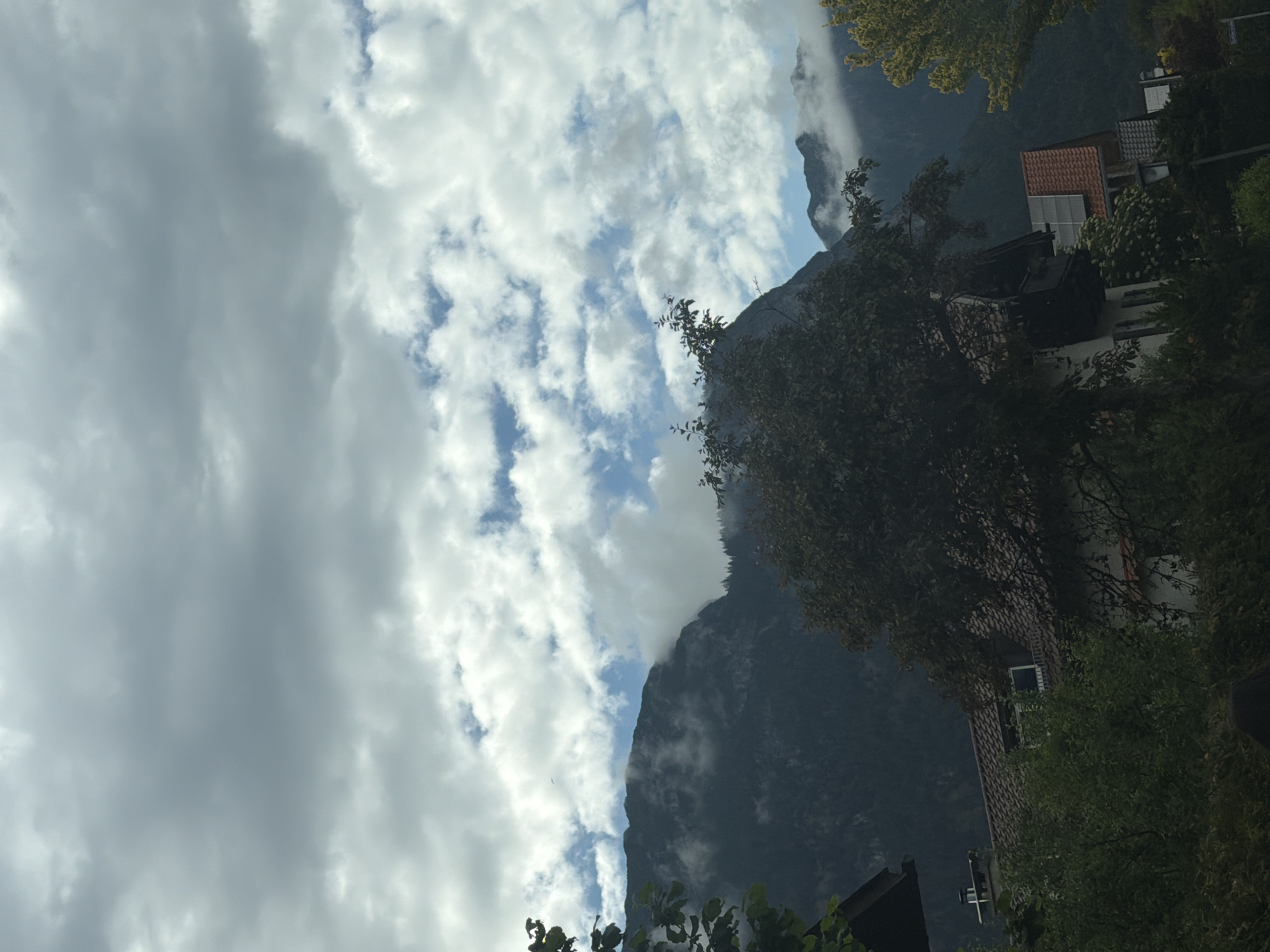 The image shows a mountain range with a partly cloudy sky. In the foreground, there are trees and houses partially visible. The scene appears to be in a mountainous region with residential areas. The image is oriented sideways, showing a landscape view.