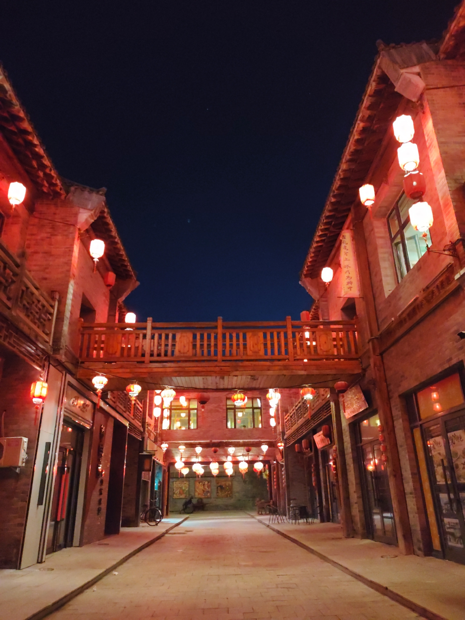The image captures a night scene on a street in Lindian, China, likely a historical or culturally significant area. Red lanterns are prominently displayed, illuminating the brick buildings and a wooden walkway connecting two buildings overhead. The architecture appears traditional, with tiled roofs and decorative wooden elements. The street is paved with cobblestones or similar material. There are no people visible in the image, but the presence of a bicycle and some chairs suggests that the area is frequented. The sky above is dark, indicating it is nighttime. The overall atmosphere is festive and traditional, enhanced by the warm glow of the lanterns.
